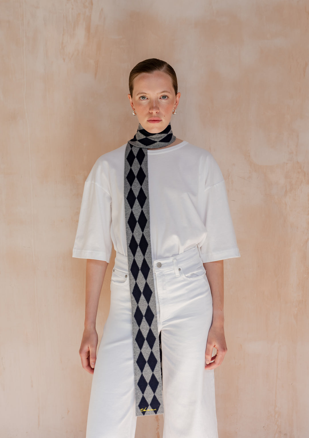 A model wearing TBCo Merino Wool Skinny Scarf in Charcoal Harlequin, featuring grey and dark-navy diamond pattern, over a white T-shirt and high-waisted jeans.