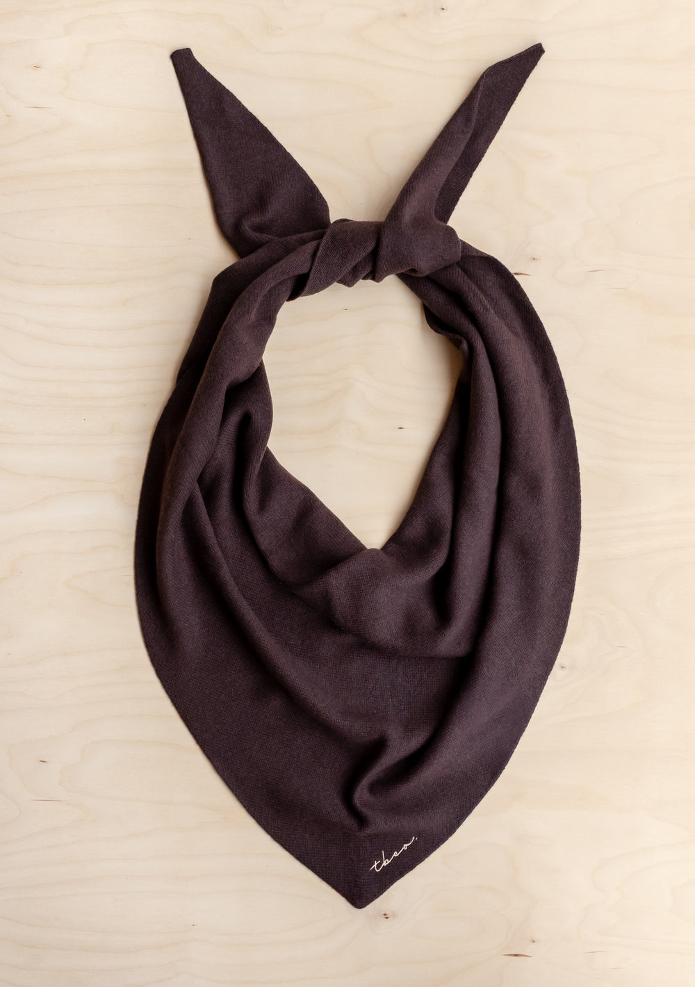 Dark-brown TBCo Large Merino Wool Triangle Scarf, knotted corners, soft drape, embroidered logo, laid on a light wood surface.