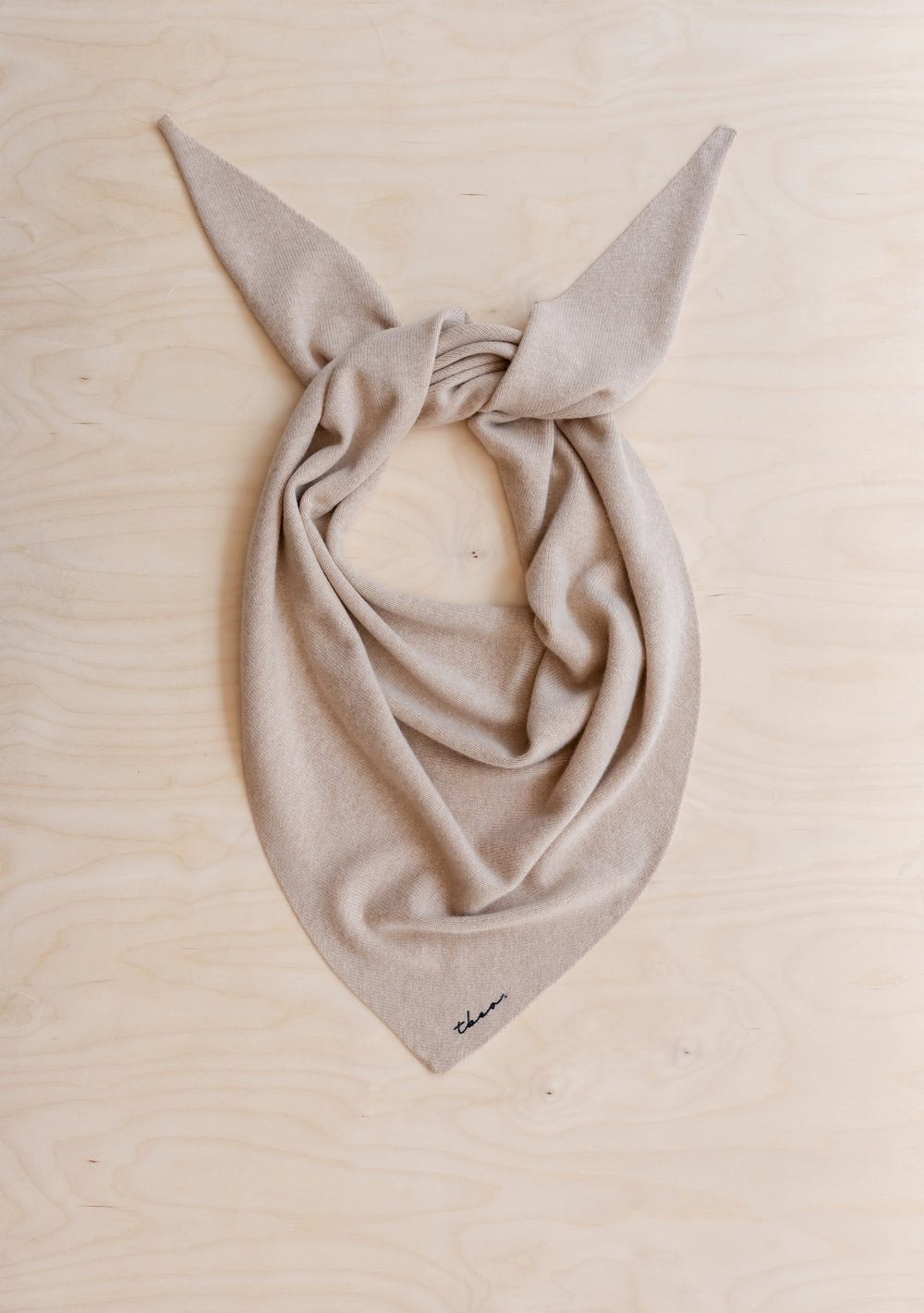 TBCo Large Merino Wool Triangle Scarf in Oatmeal, light-beige, folded, knotted, with TBCo embroidered logo near the bottom, laid on wooden surface.