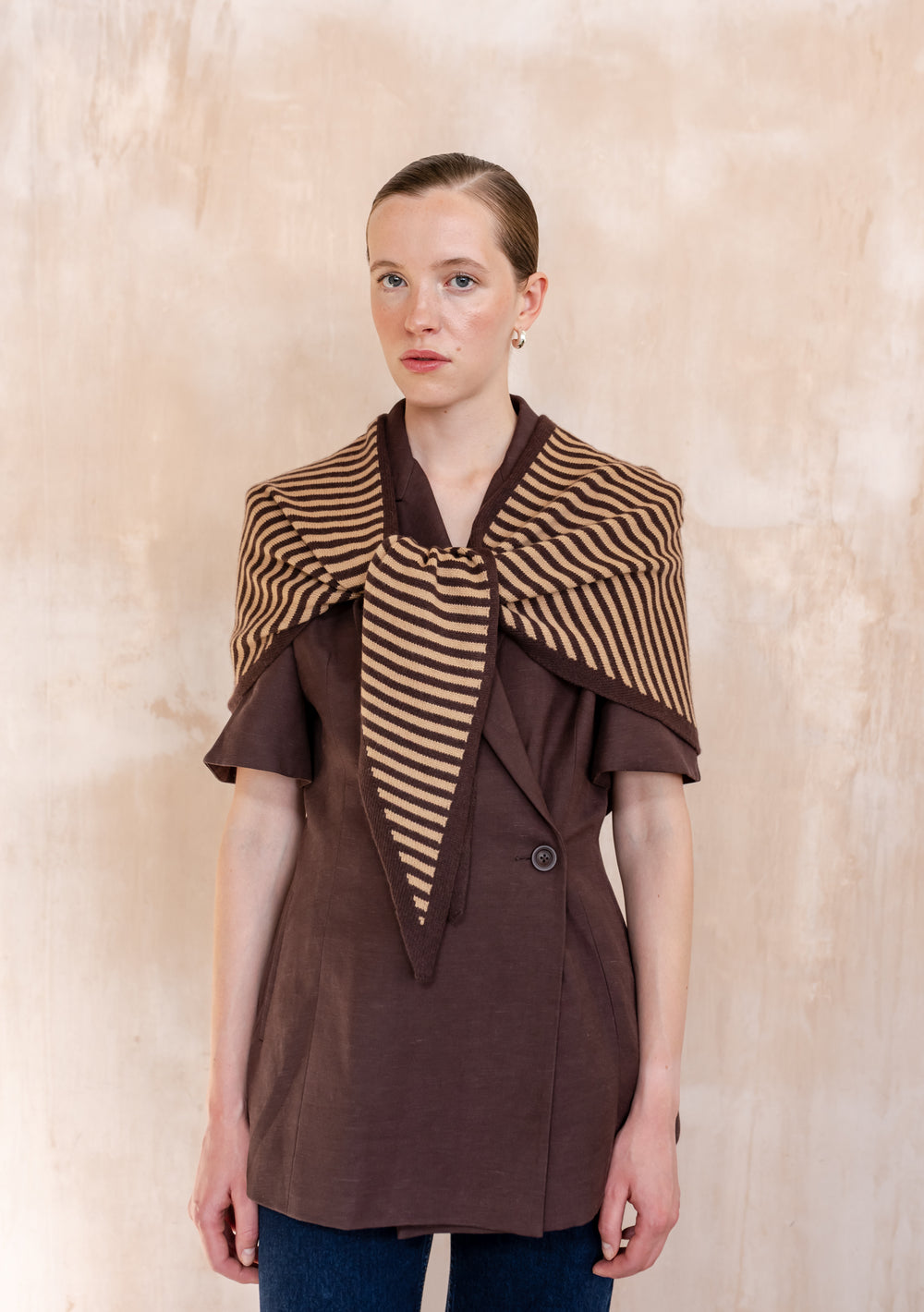 Young woman models the TBCo Large Merino Wool Triangle Scarf in Camel & Brown Stripe, draped over a brown jacket, showcasing its striped design and versatile style.