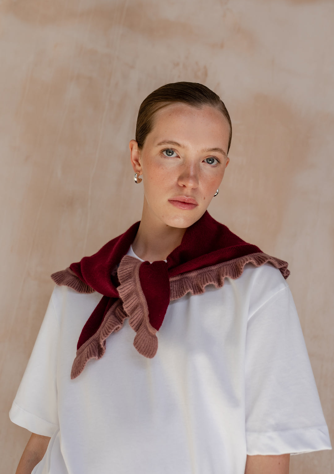 Young woman wearing TBCo Merino Wool Triangle Scarf in Burgundy Ruffle, featuring playful rose trim, draped over white T-shirt, showcasing elegant silver hoop earrings.