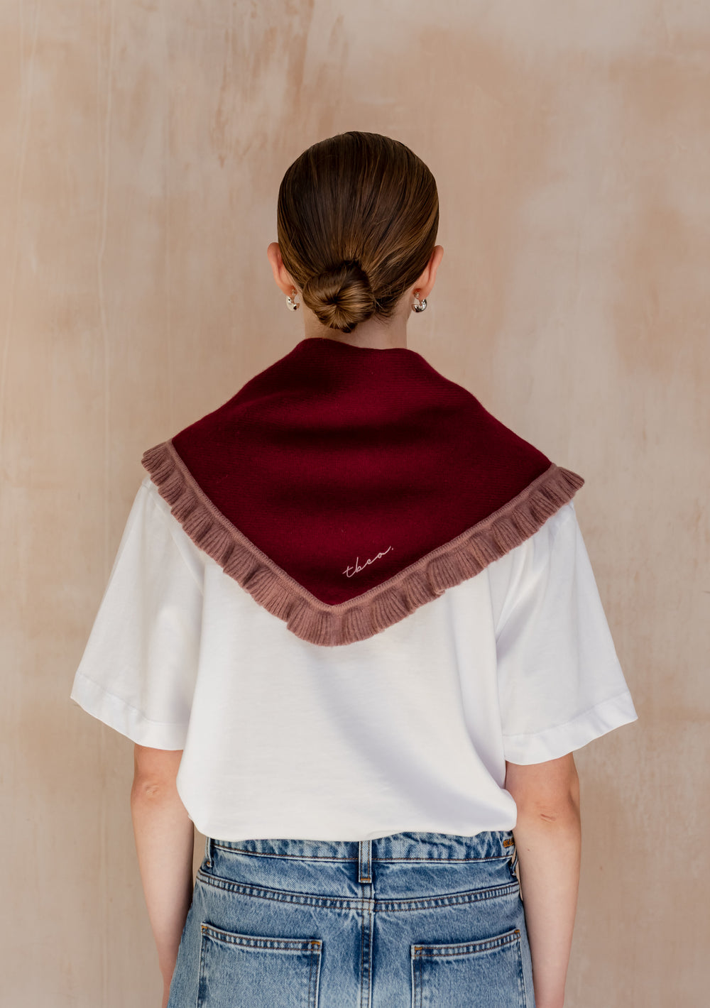 Person wearing TBCo Merino Wool Triangle Scarf in Burgundy Ruffle over white T-shirt and jeans, showcasing its burgundy tone and taupe ruffle trim against a beige wall.