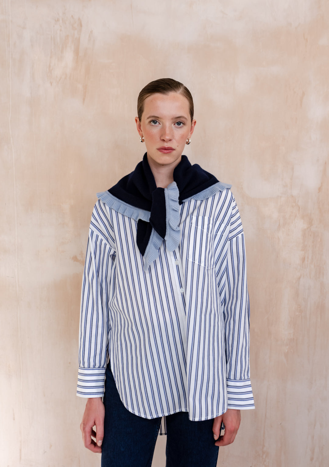 Young woman models the TBCo Merino Wool Triangle Scarf in Navy Ruffle, draped stylishly over a striped shirt, showcasing its light-blue ruffle trim and elegant design.