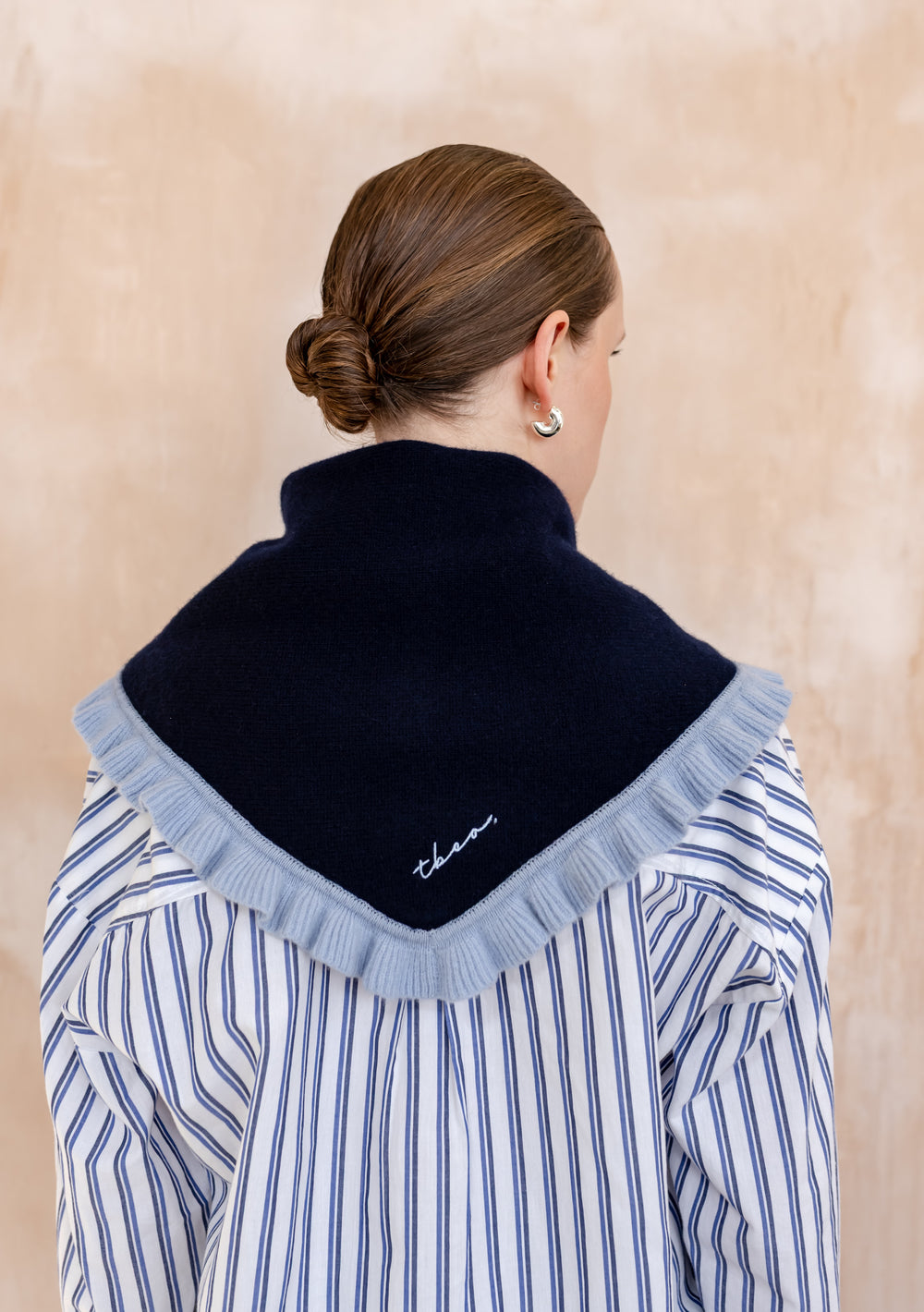 A woman wears the TBCo Merino Wool Triangle Scarf in Navy Ruffle, draped over her shoulders, showcasing its light-blue ruffle trim and embroidered logo.