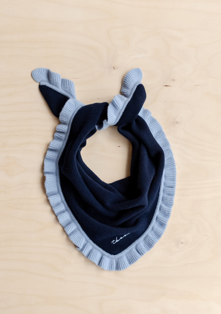 TBCo Merino Wool Triangle Scarf in Navy Ruffle featuring deep navy knit with pale blue ruffle trim, knotted corners, and embroidered logo, laid flat on wood surface