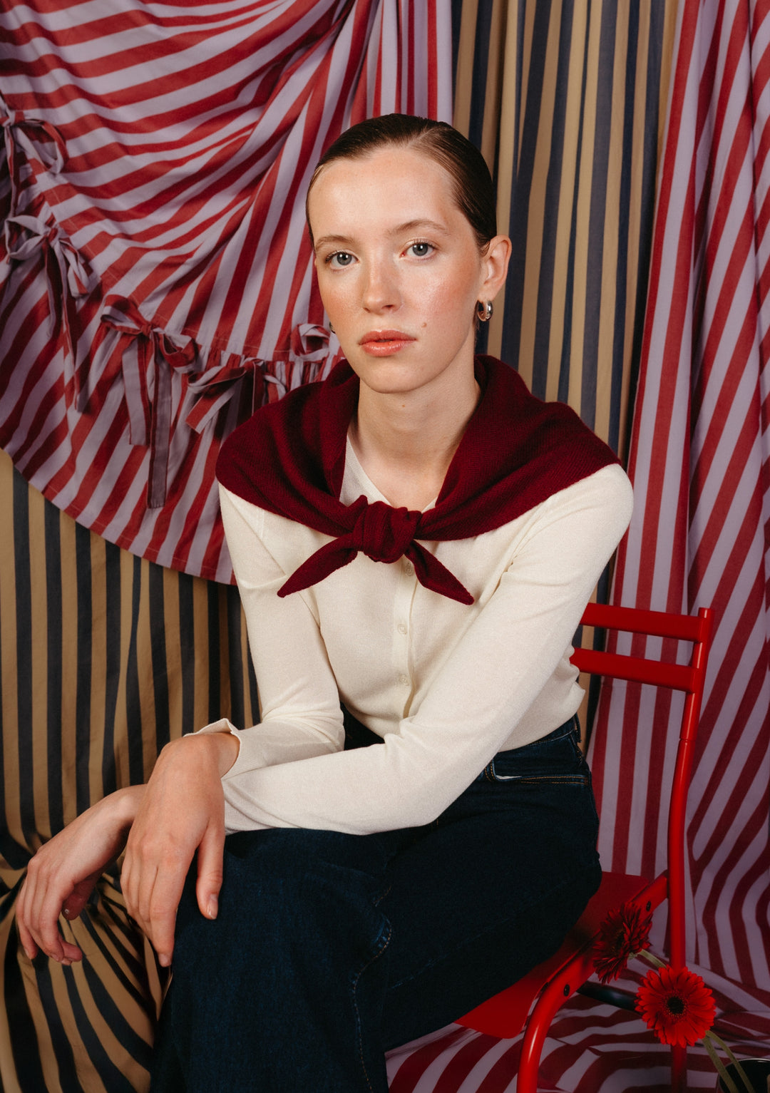 Young woman models the TBCo Merino Wool Triangle Scarf in Burgundy, styled over her shoulders, complementing her cream top and dark jeans against a striped backdrop.