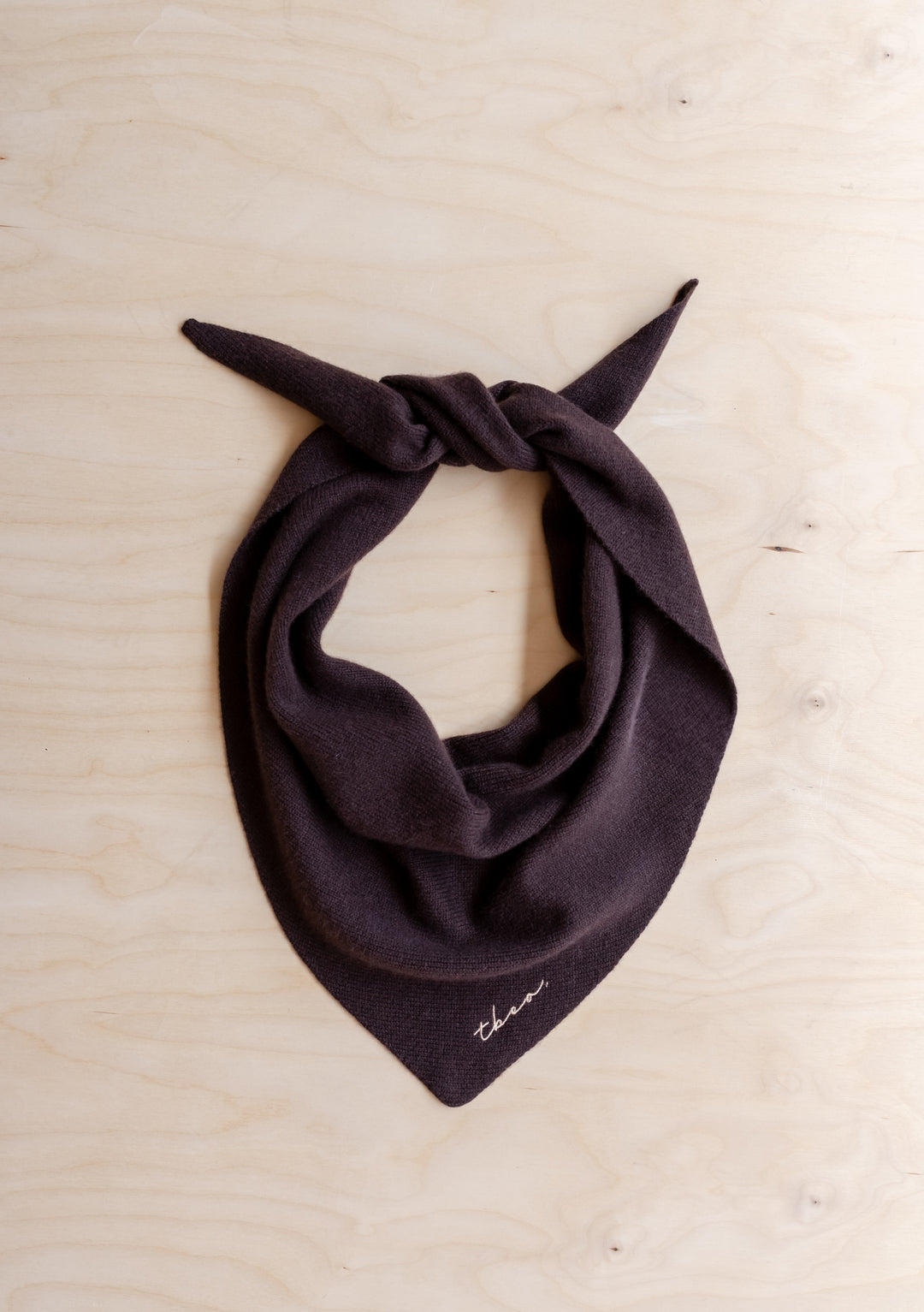 TBCo Merino Wool Triangle Scarf in Brown, featuring a knotted design and embroidered logo, laid on wood surface, showcasing its softness and versatile styling.