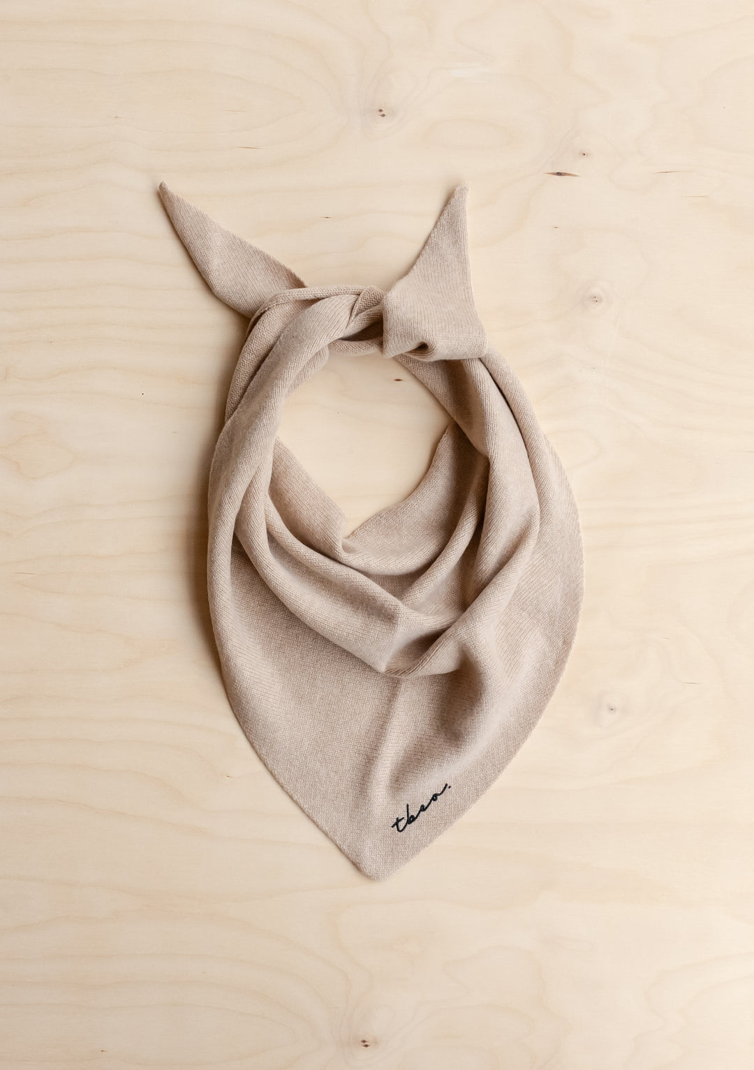 TBCo Merino Wool Triangle Scarf in Oatmeal, featuring taupe knit, black embroidery, and knotted ends, laid flat on wooden surface.