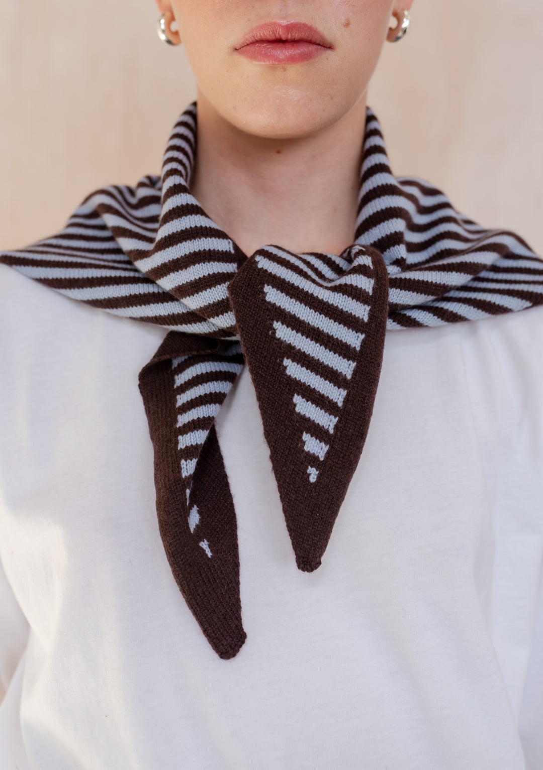 A person wears a TBCo Merino Wool Triangle Scarf in Blue & Brown Stripe with pointed ends, styled over a white top, showcasing small hoop earrings.