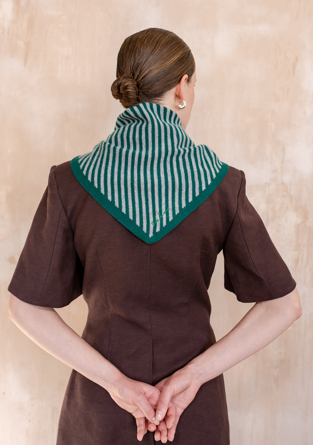 Woman wearing TBCo Merino Wool Triangle Scarf in Green & Charcoal Stripe, draped over shoulders, showcasing green and grey knitted pattern, with hands clasped behind.