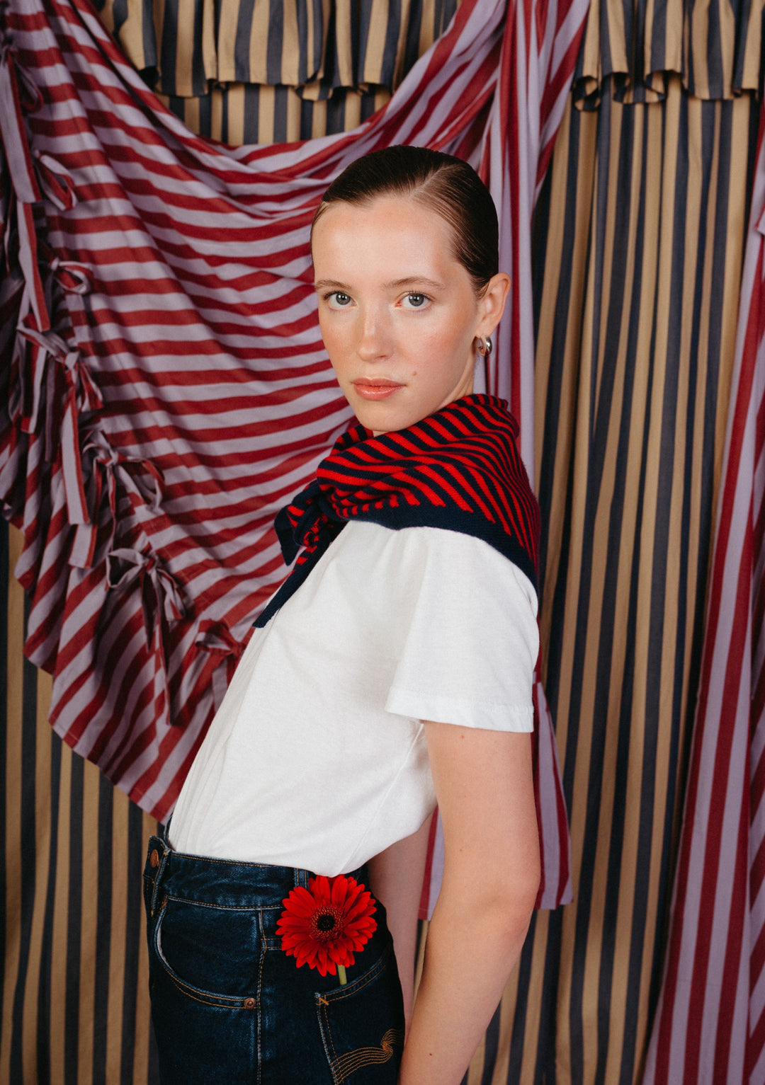 Young person models the TBCo Merino Wool Triangle Scarf in Navy & Red Stripe, casually draped over shoulders, enhancing a white T-shirt and denim jeans ensemble.