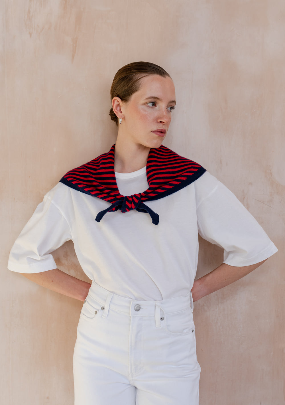 A young person wears a TBCo Merino Wool Triangle Scarf in Navy & Red Stripe, draped over shoulders, paired with a white outfit, showcasing casual elegance.