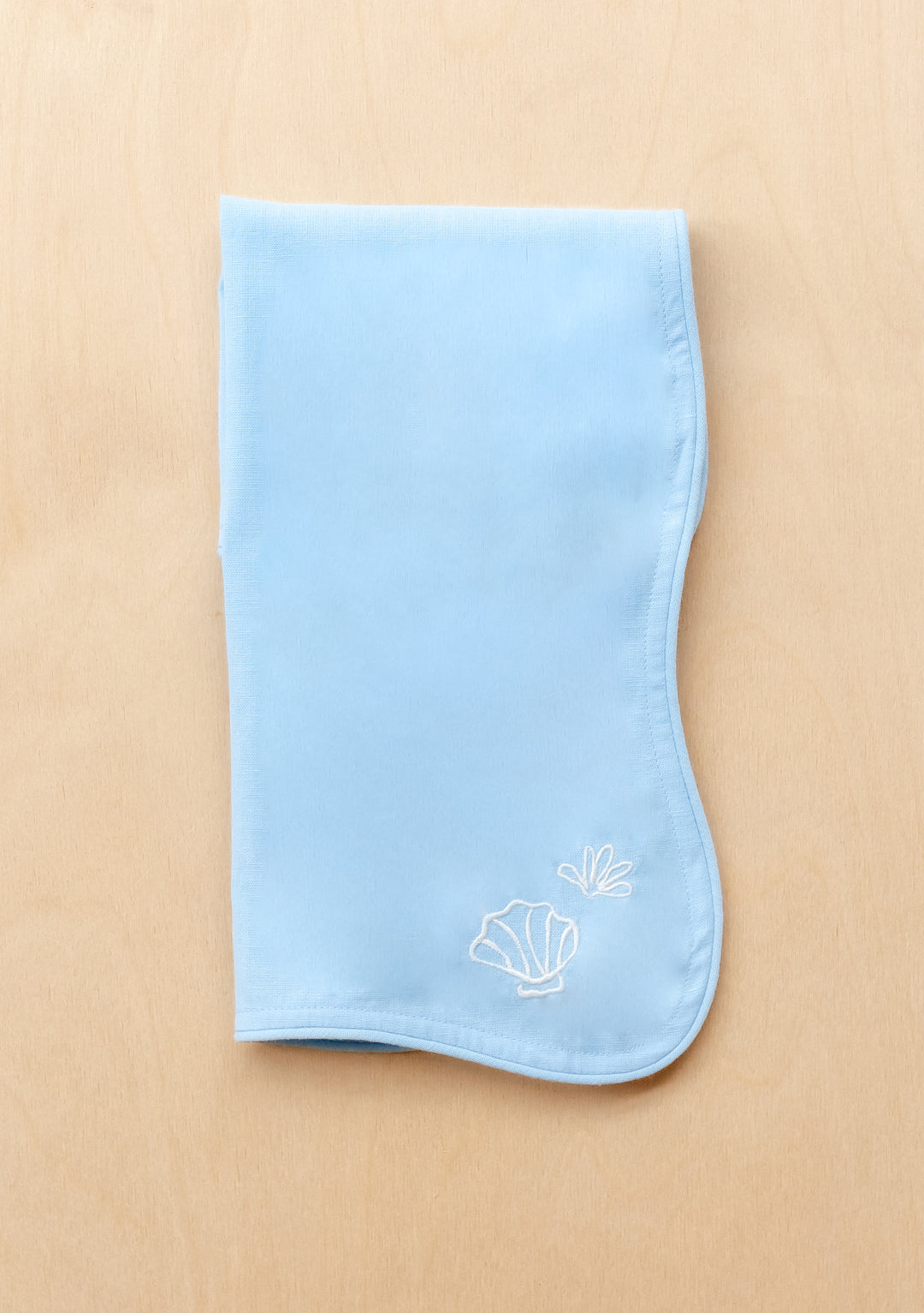 TBCo Cotton & Linen Napkins in light blue with white embroidered seashells and scalloped edges on beige surface