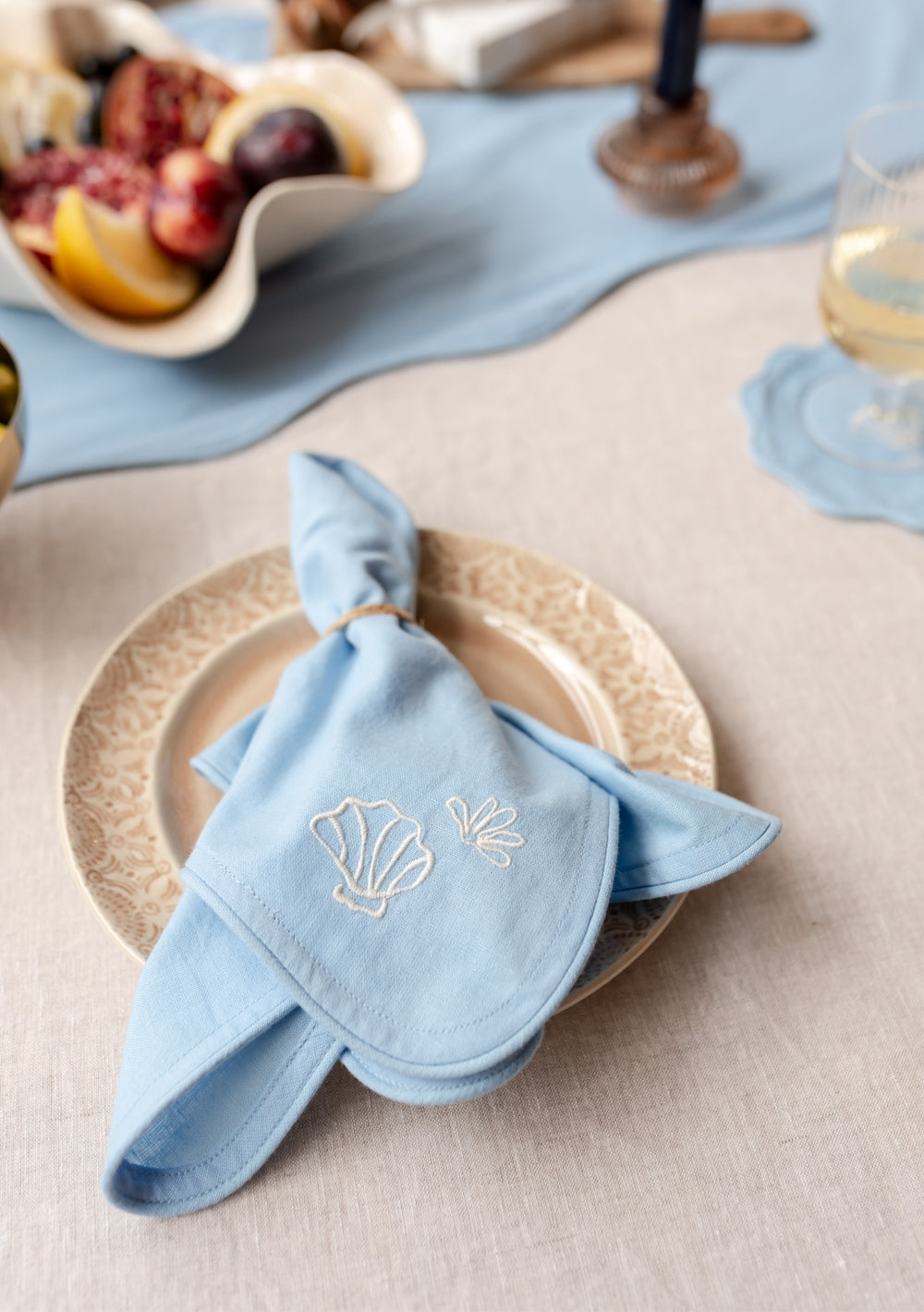 TBCo Cotton & Linen Napkins in light blue with embroidered seashell design, elegantly folded on cream plate at table setting