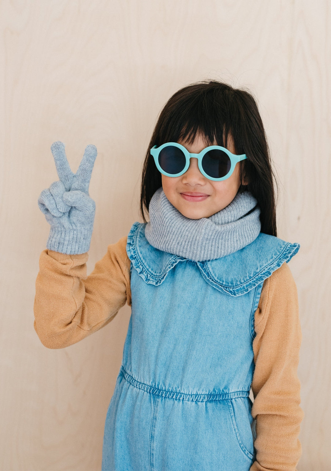 Child wearing TBCo Merino Wool Kids Neck Warmer in Light Grey Melange with denim dungarees, showing peace sign gesture