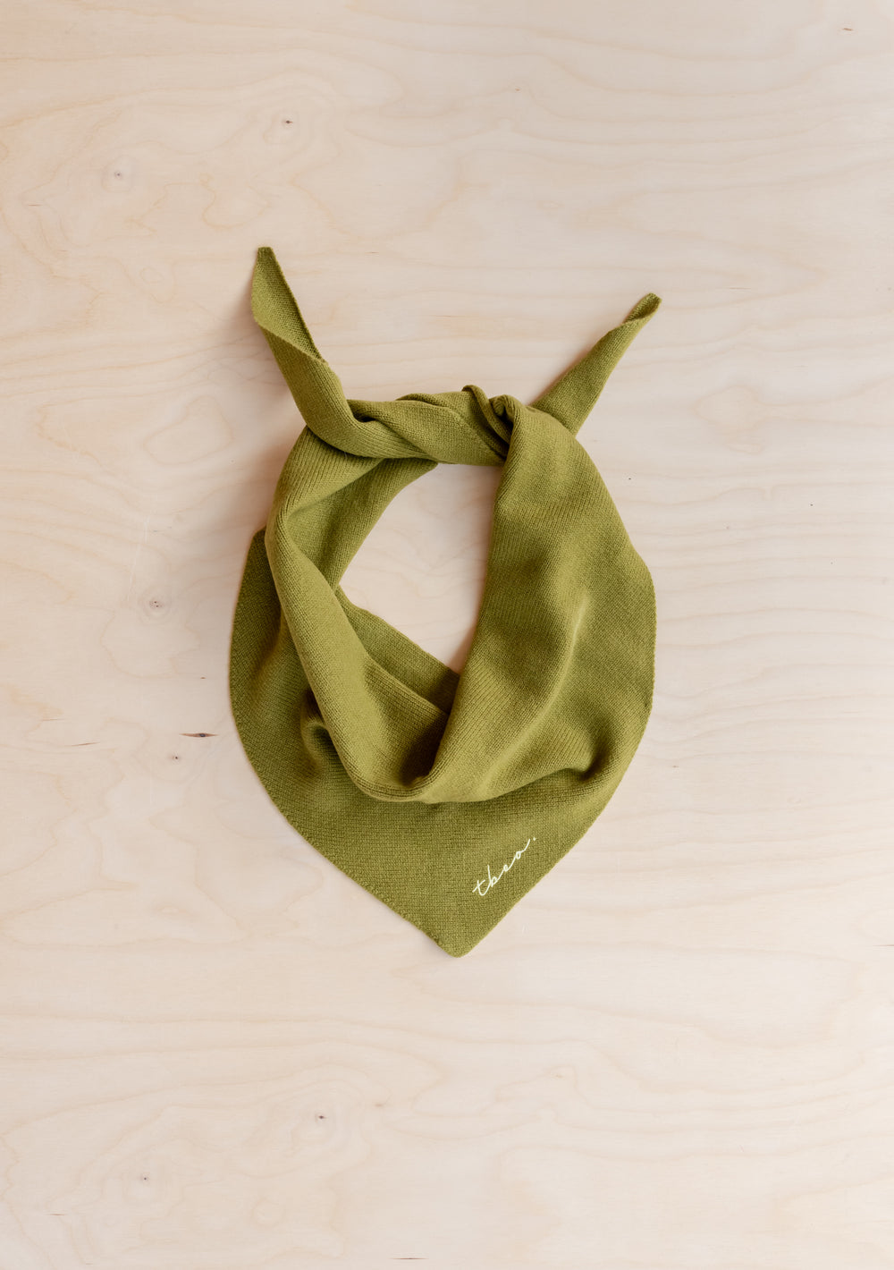 TBCo Merino Triangle Scarf in Olive, crafted from soft wool, styled in a knotted position on wooden surface with yellow logo detail