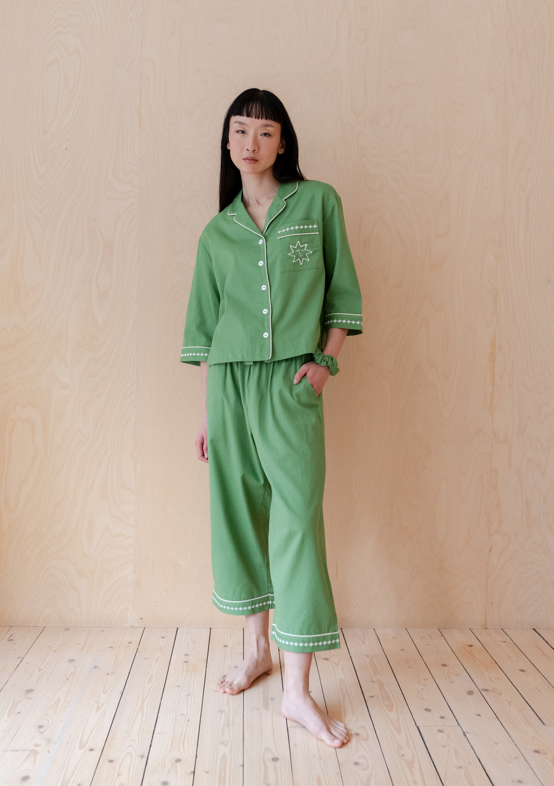 Green cotton and linen TBCo pyjama set with embroidered sun motif and matching scrunchie, shown on wooden floor