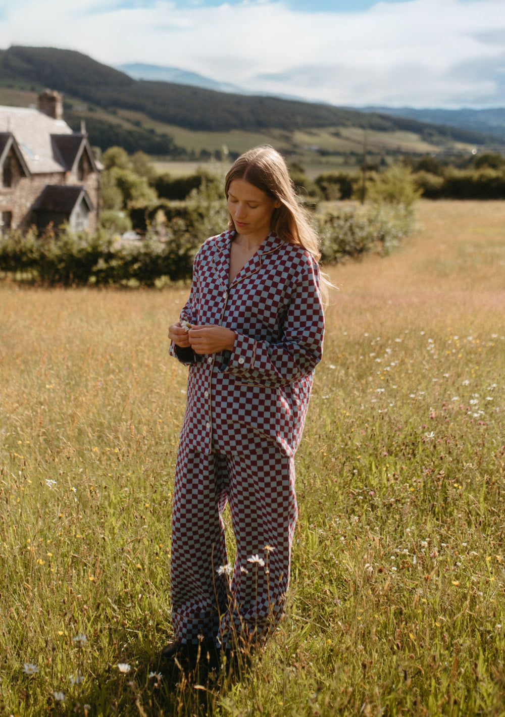 Woman wearing TBCo Cotton Pyjamas in Blue Checkerboard standing in a sunlit meadow with hills in the background.