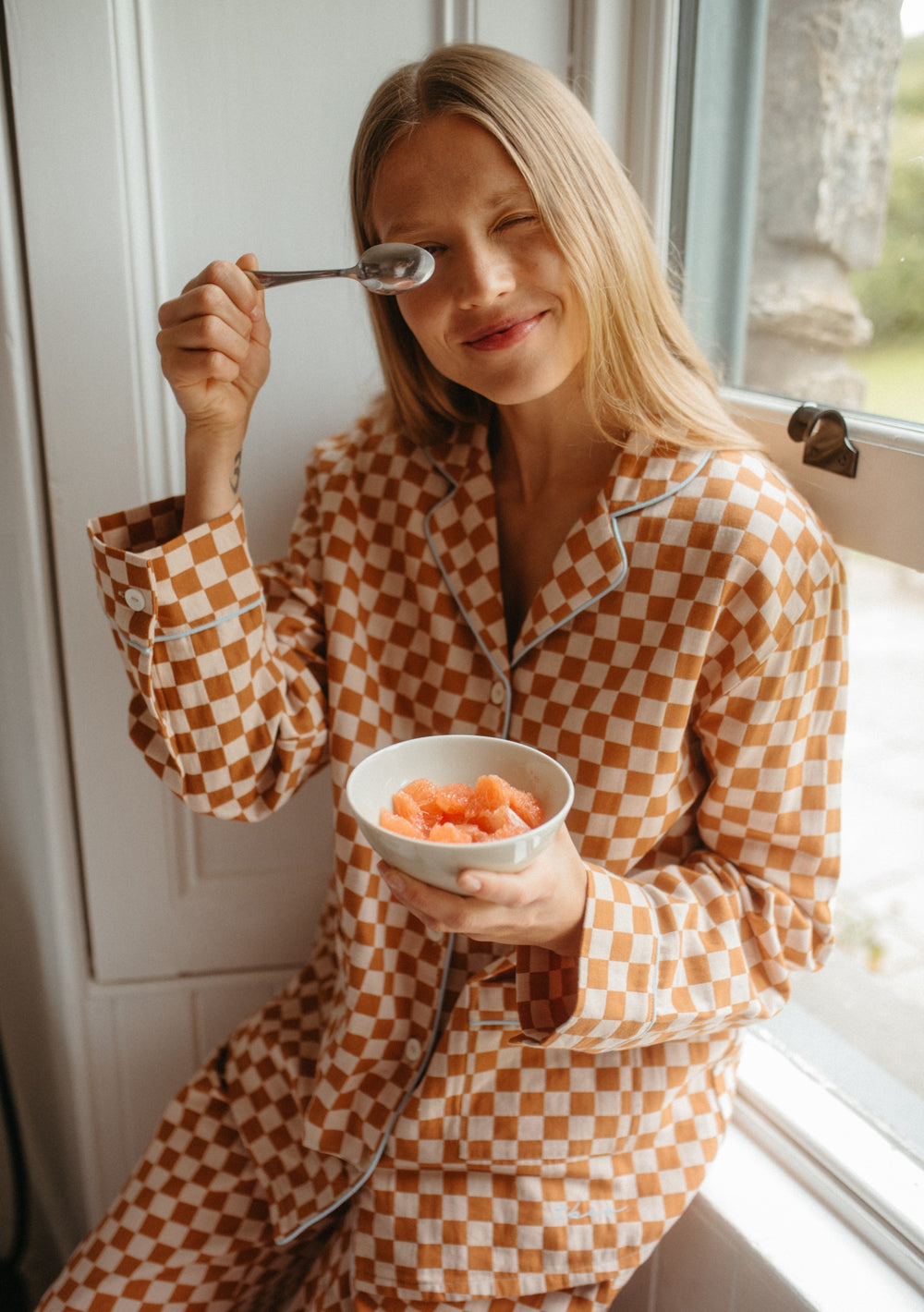 Woman wearing TBCo Cotton Pyjamas in Honey Checkerboard holding a bowl of fruit by the window.