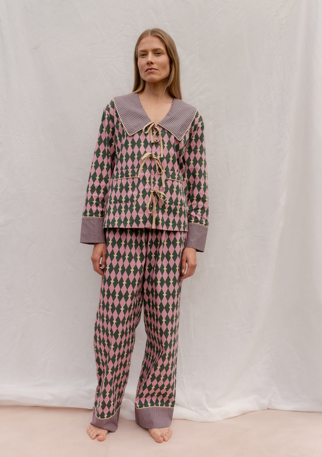 Model wearing TBCo Cotton Pyjamas in Pink Harlequin with a striped collar, camel bows, and breathable cotton fabric.