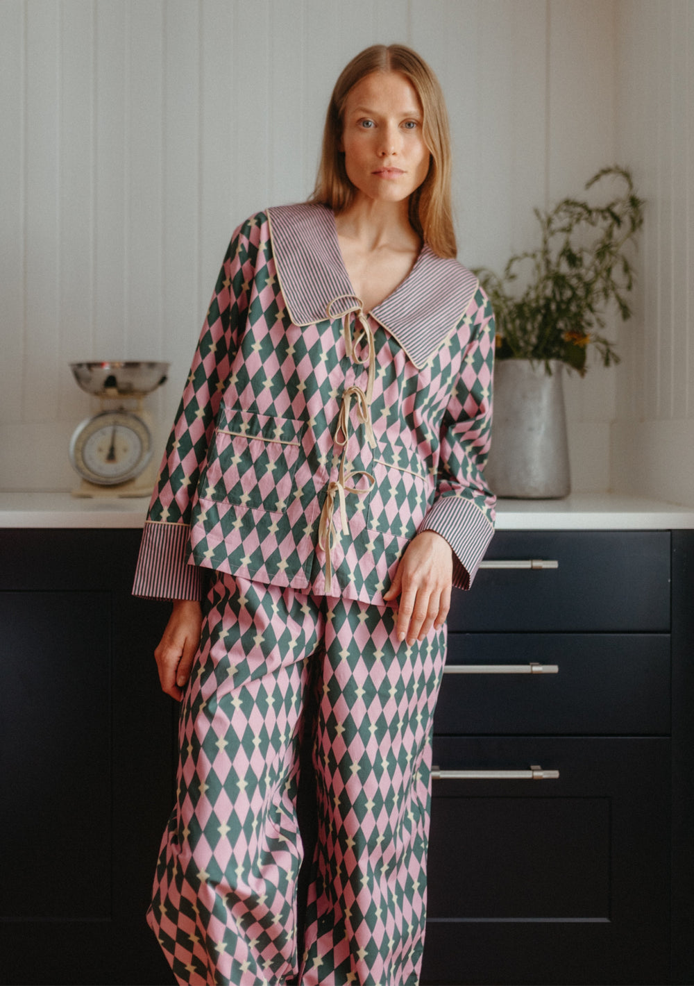 Woman wearing TBCo Cotton Pyjamas in Pink Harlequin with bow ties and striped collar in a kitchen setting.
