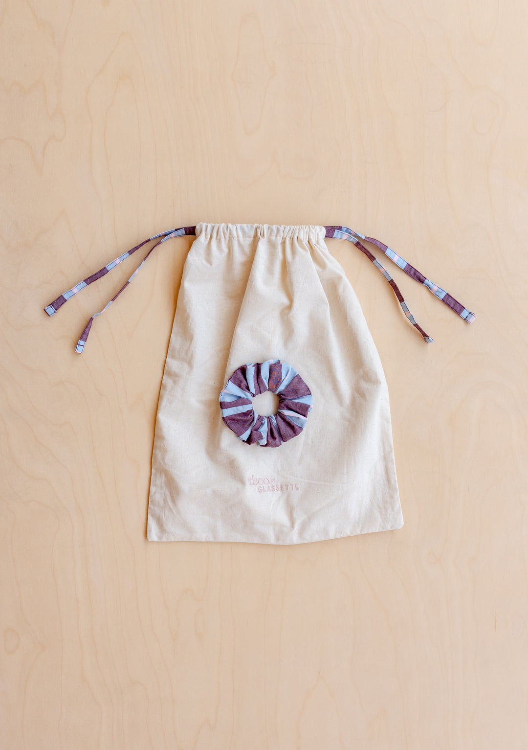TBCo x Glassette Cotton Pyjamas in Berry Stripe with a striped scrunchie on a natural-canvas pouch, featuring purple-and-blue ties, laid flat on wood.