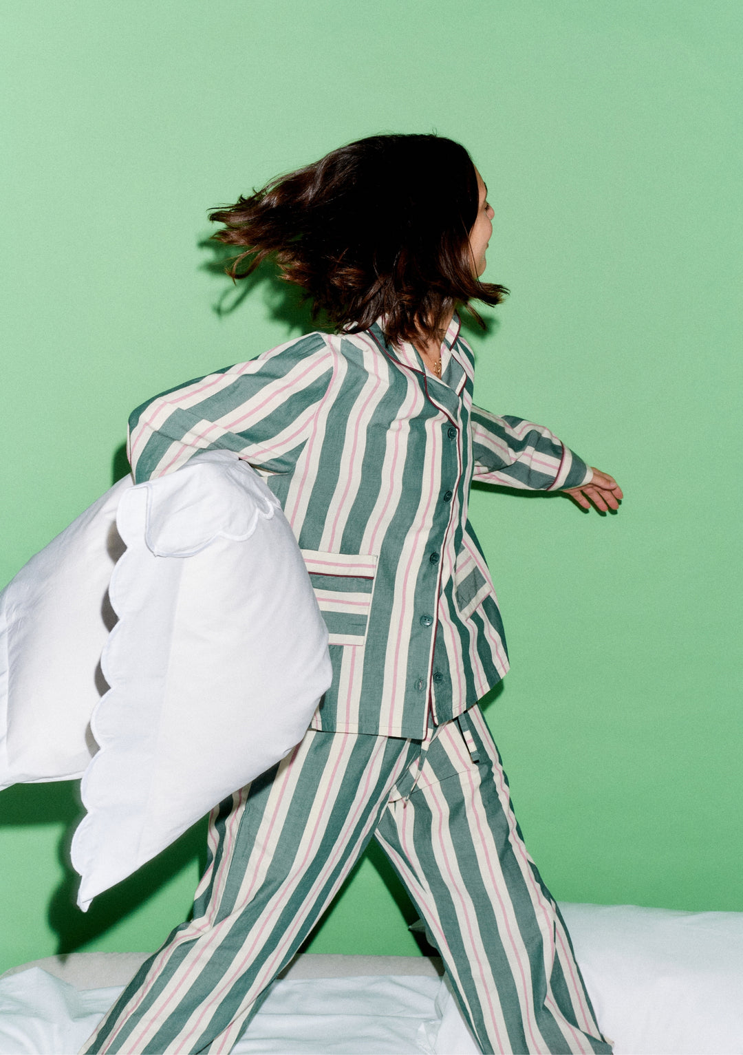 Person in motion wearing TBCo x Glassette Cotton Pyjamas in Green Stripe, holding a white pillow, showcasing the vibrant, comfortable design against a light-green background.