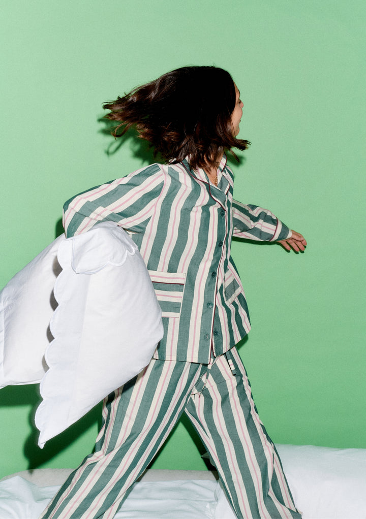 Person in motion wearing TBCo x Glassette Cotton Pyjamas in Green Stripe, holding a white pillow, showcasing the vibrant, comfortable design against a light-green background.
