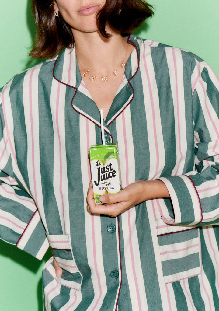 Person in TBCo x Glassette Cotton Pyjamas in Green Stripe, holding a green-and-white juice carton with a straw, showcasing cozy loungewear style.