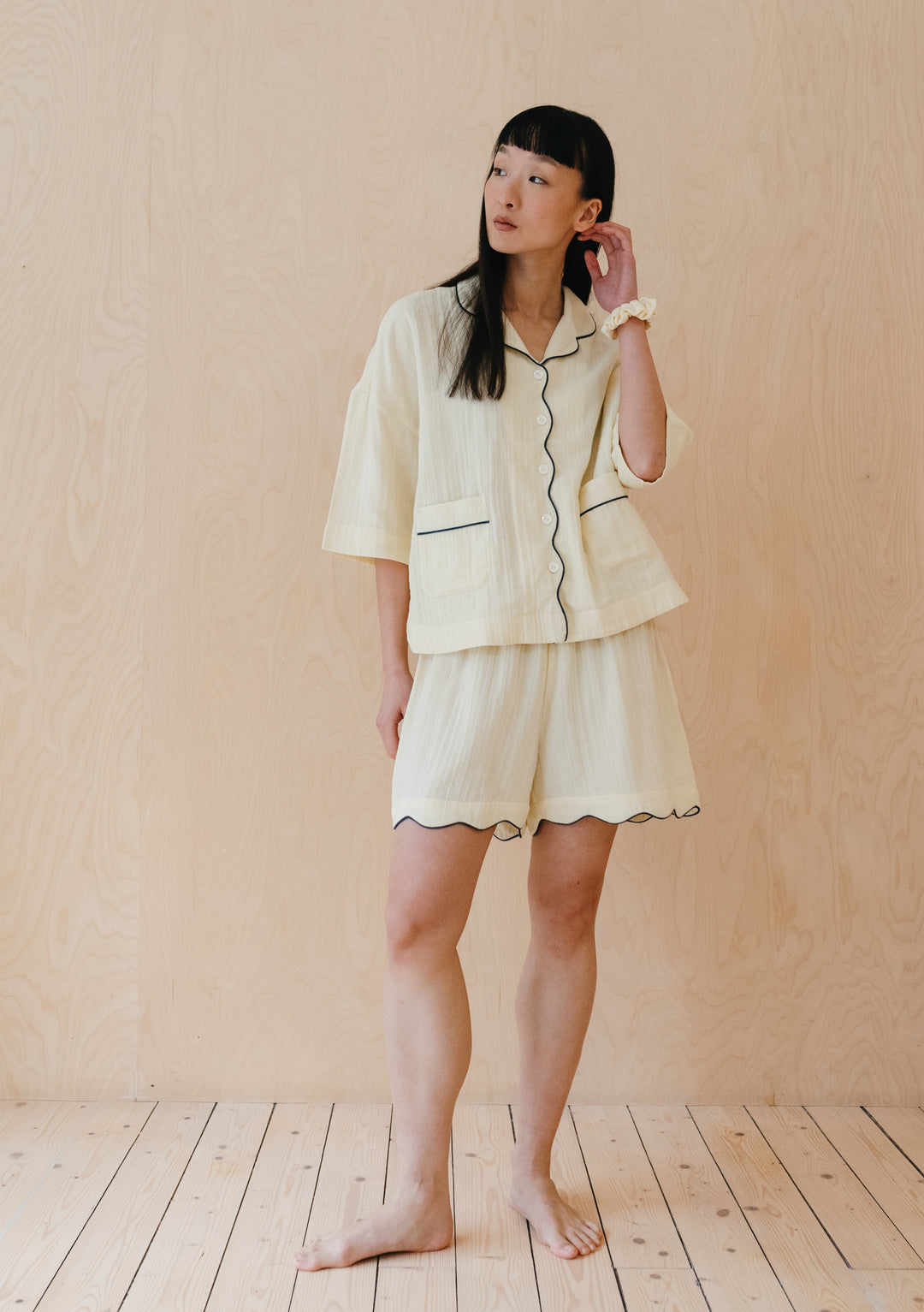 A person wearing TBCo Cotton Pyjamas in Buttermilk with navy piping, featuring scalloped shorts and short-sleeve top against wooden backdrop