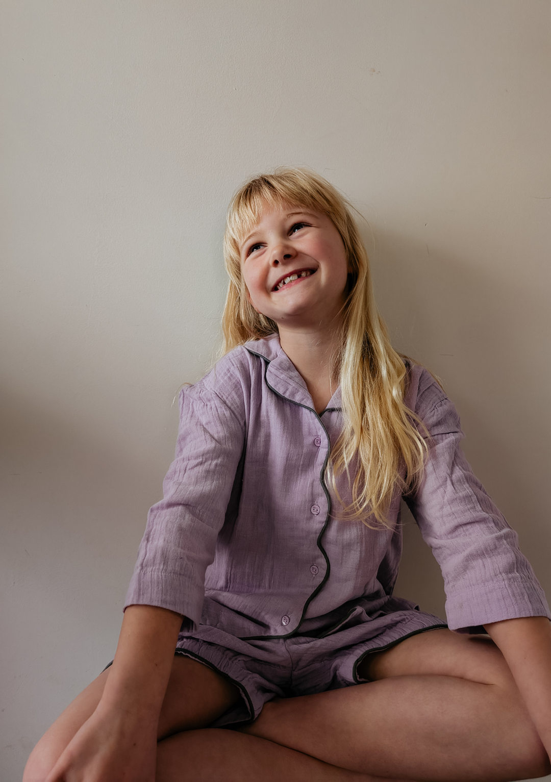 Child sitting wearing TBCo Cotton Kids Pyjamas in Lilac with button front and contrast trim on lightweight fabric