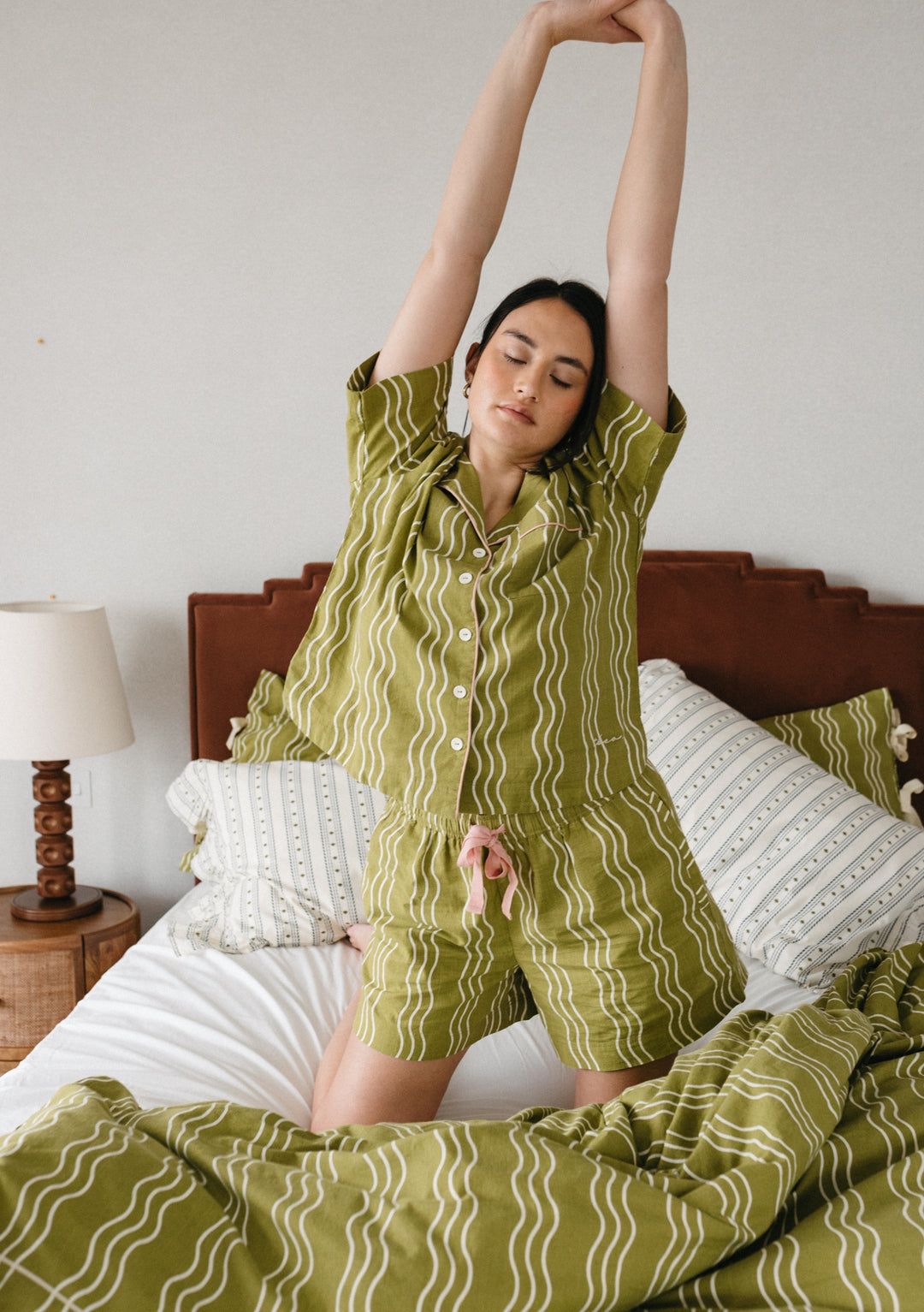 Person stretching in TBCo Cotton Pyjamas in Olive Wave pattern, showing comfortable fit on bed with matching bedding