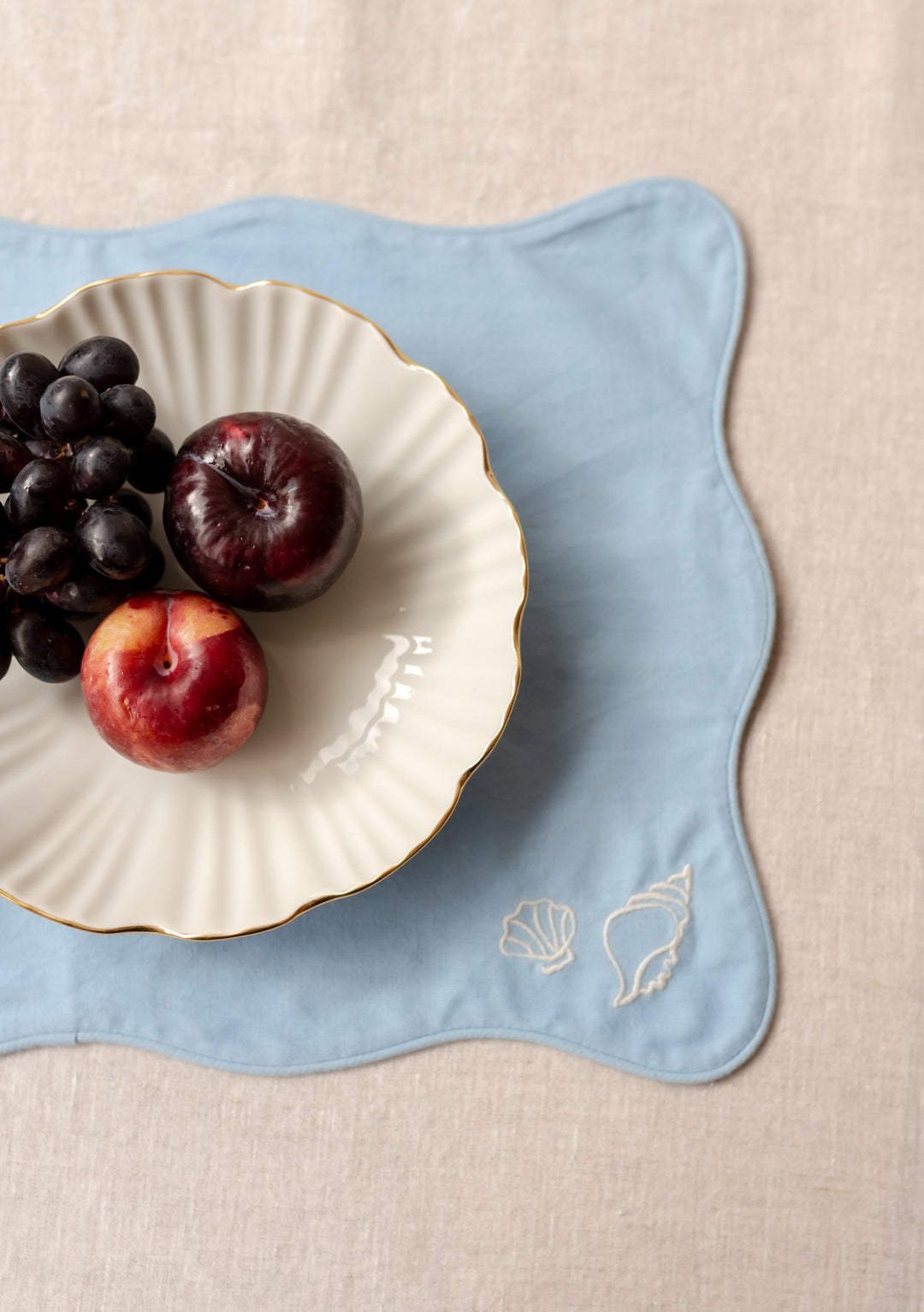 TBCo Cotton & Linen Placemat in blue with scalloped edges and seashell embroidery shown with white plate holding fresh fruit