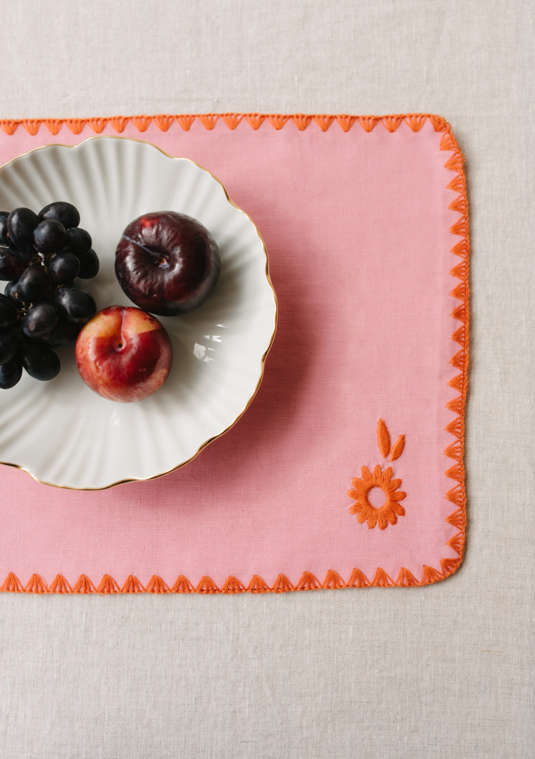 Pink TBCo cotton and linen placemat with orange embroidered edges and floral detail, showcasing a white plate with fresh fruit