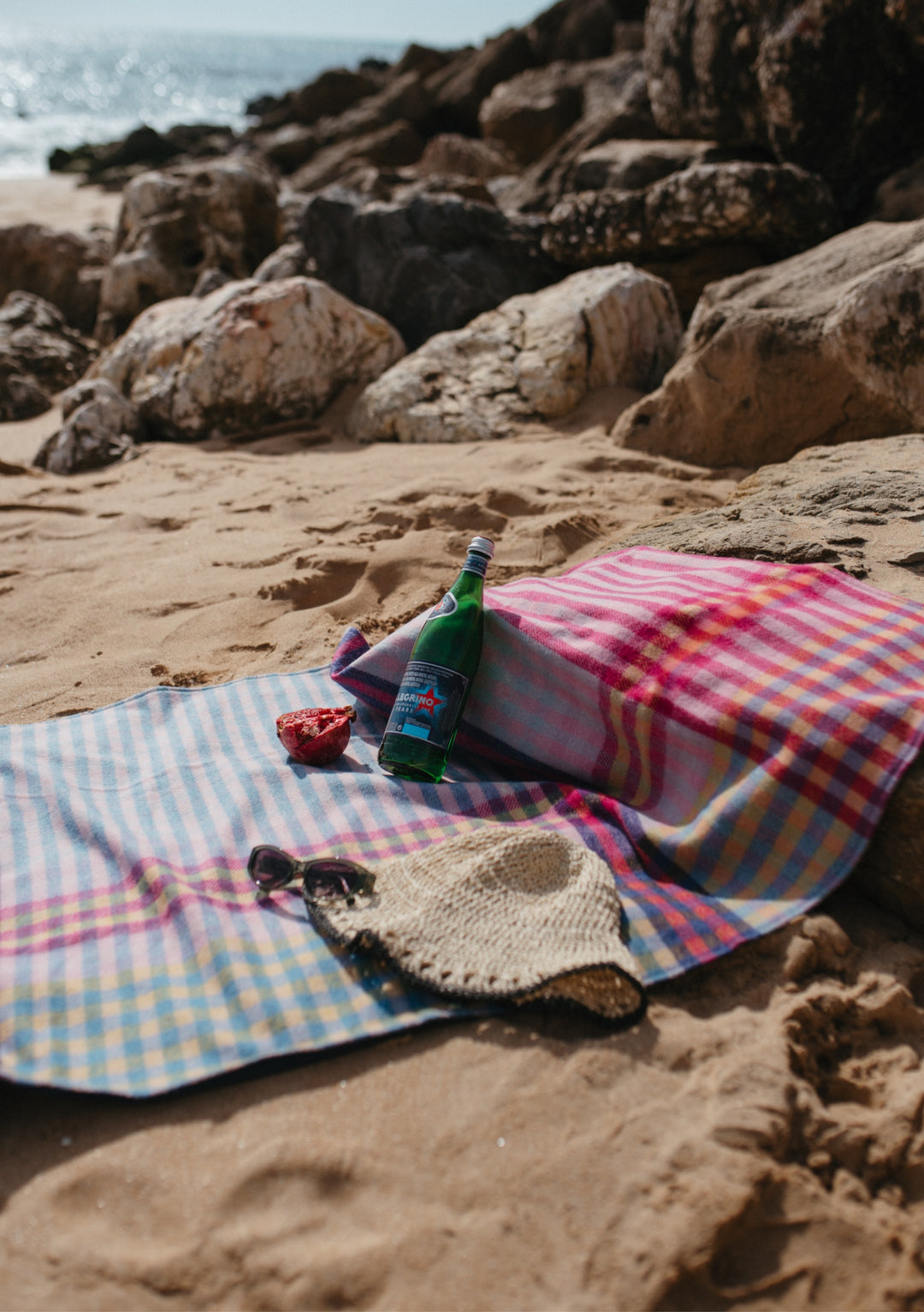 TBCo recycled wool pink gingham picnic blanket laid on sandy beach with bottle and rocks in background