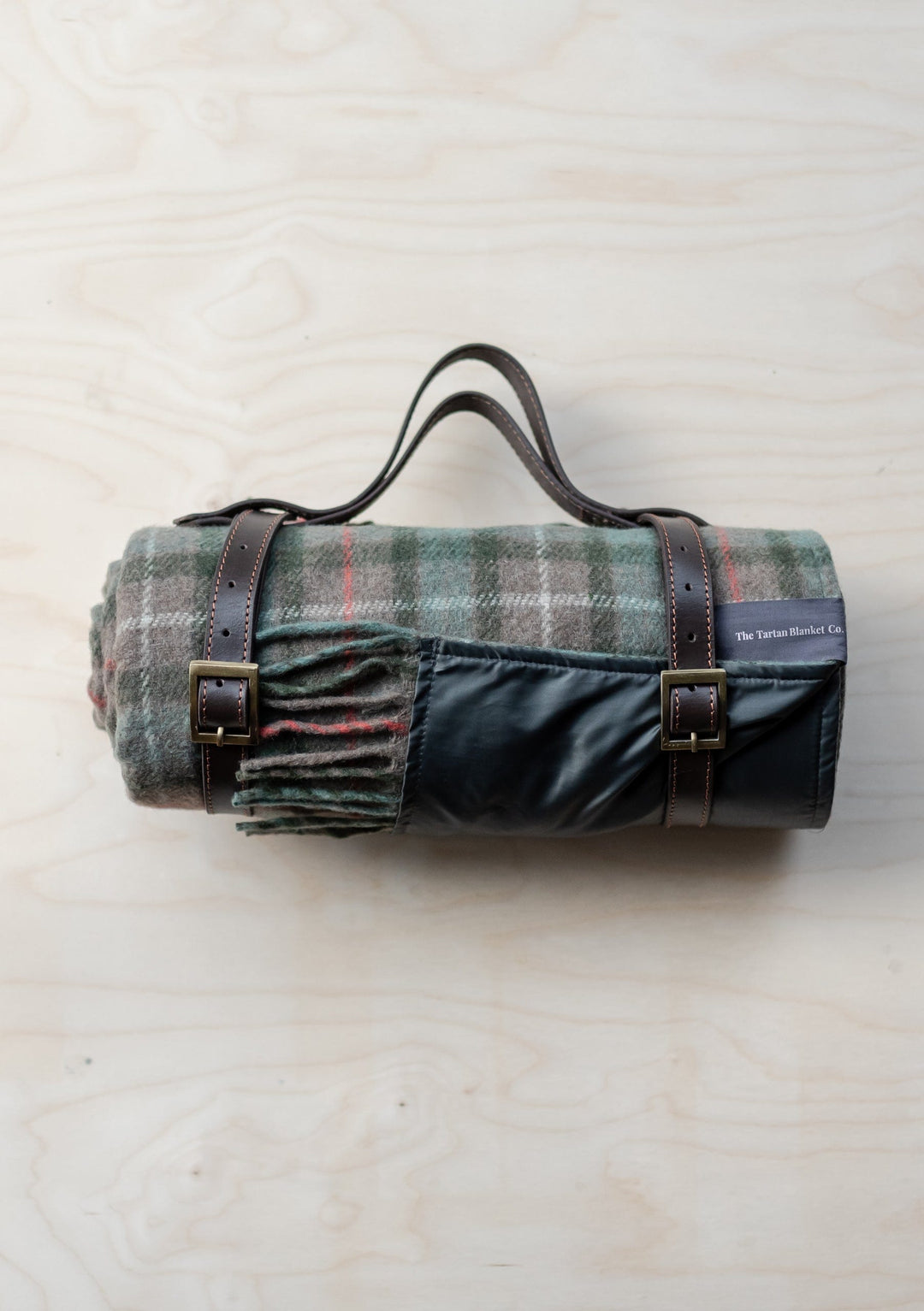 TBCo Recycled Wool Picnic Blanket in Fraser Hunting Weathered Tartan, rolled with leather carrier straps on wooden surface