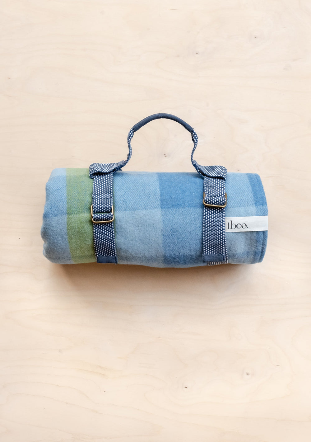 TBCo Recycled Wool Picnic Blanket in blue and green gingham pattern, rolled with grey carrying straps and brand label