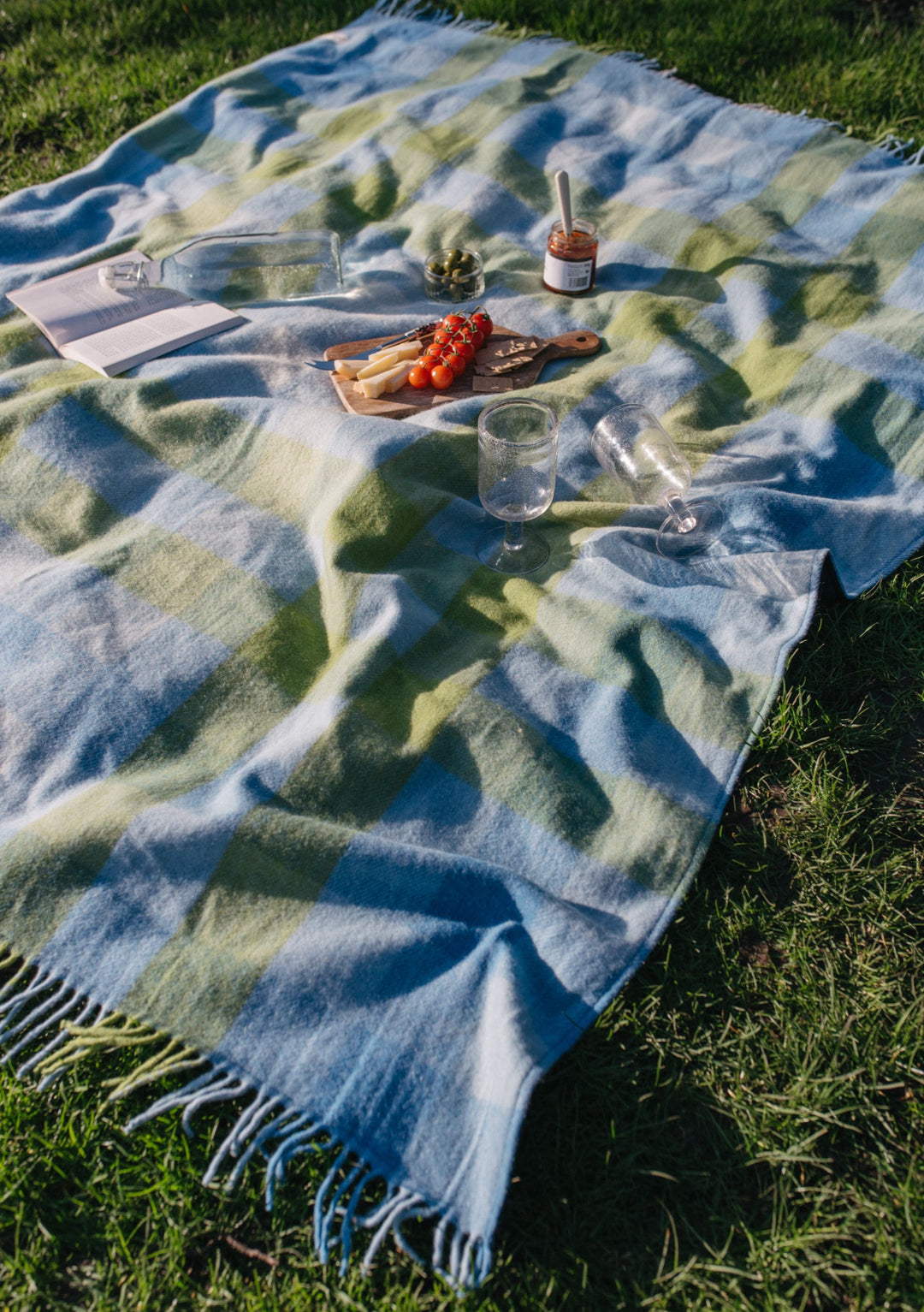 TBCo Recycled Wool Picnic Blanket in blue gradient gingham pattern laid on grass with picnic spread featuring snacks and drinks