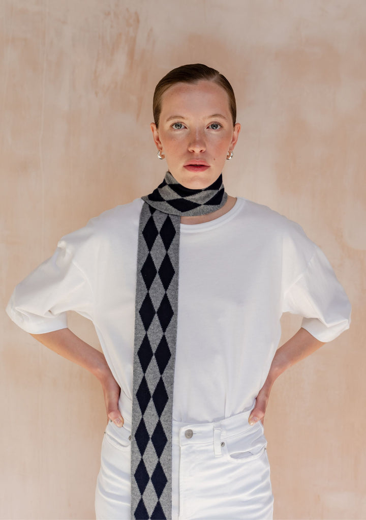 A model wearing TBCo Merino Wool Skinny Scarf in Charcoal Harlequin, featuring grey and dark-navy diamond pattern, over a white T-shirt.