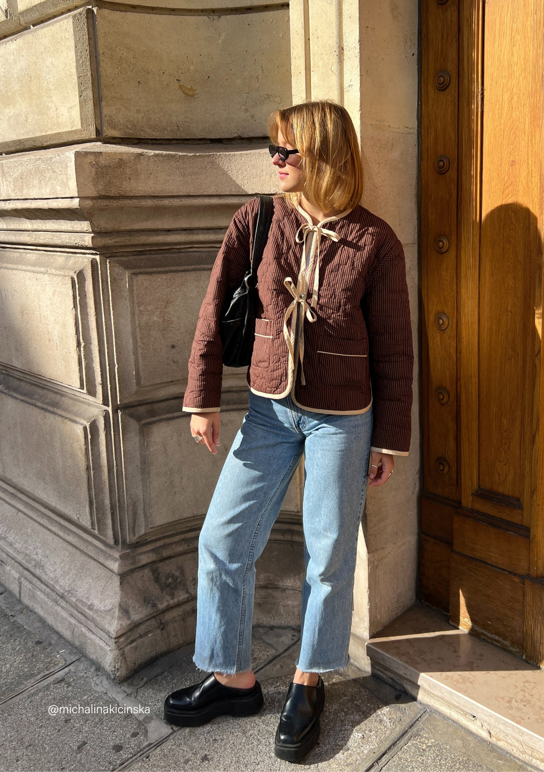 Person wearing TBCo quilted cotton jacket in reversible brown celestial stripe, styled with denim and sunglasses against stone wall