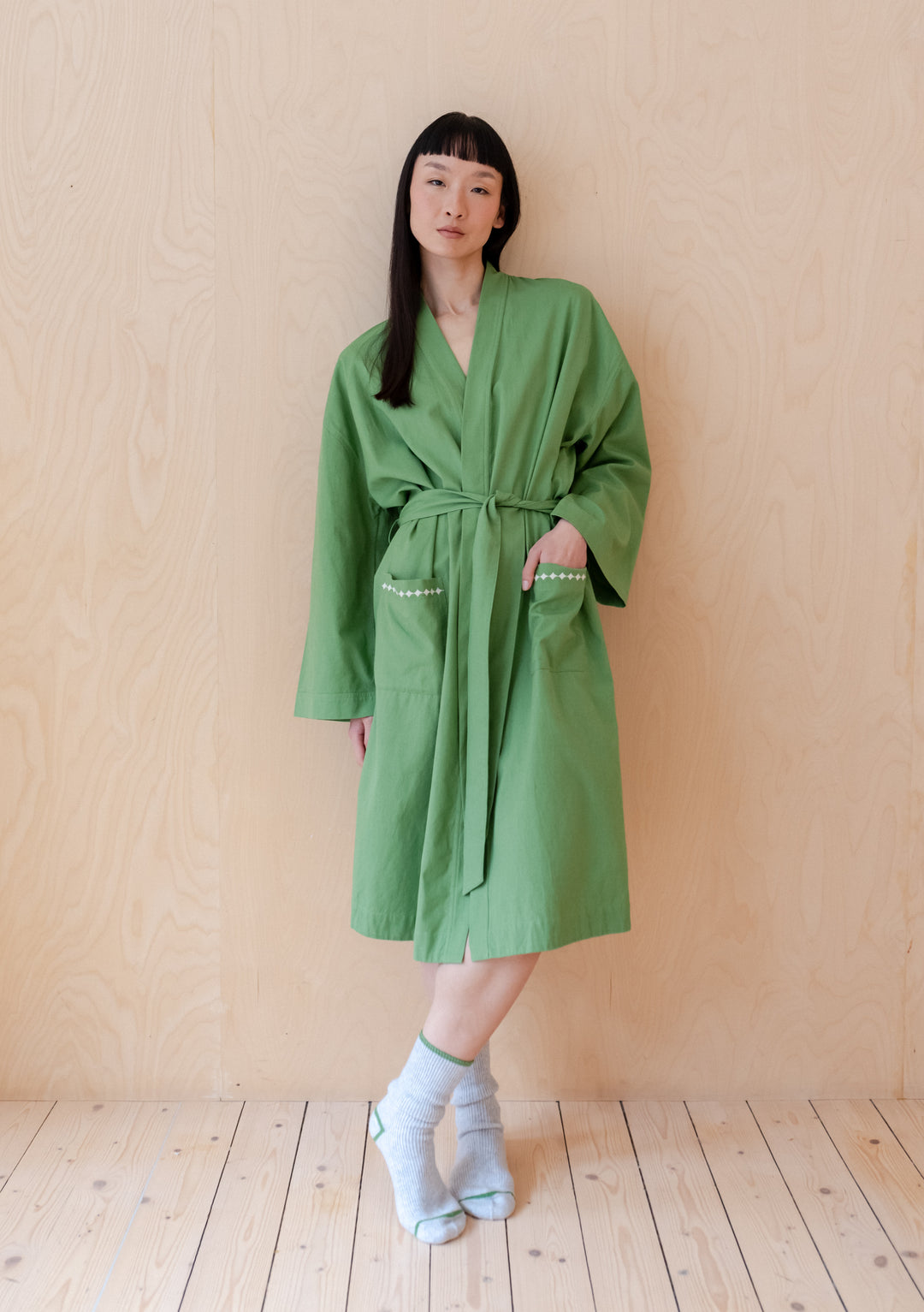 A person wearing TBCo's Cotton & Linen Robe in Green, showing relaxed fit and embroidered pockets against wooden backdrop