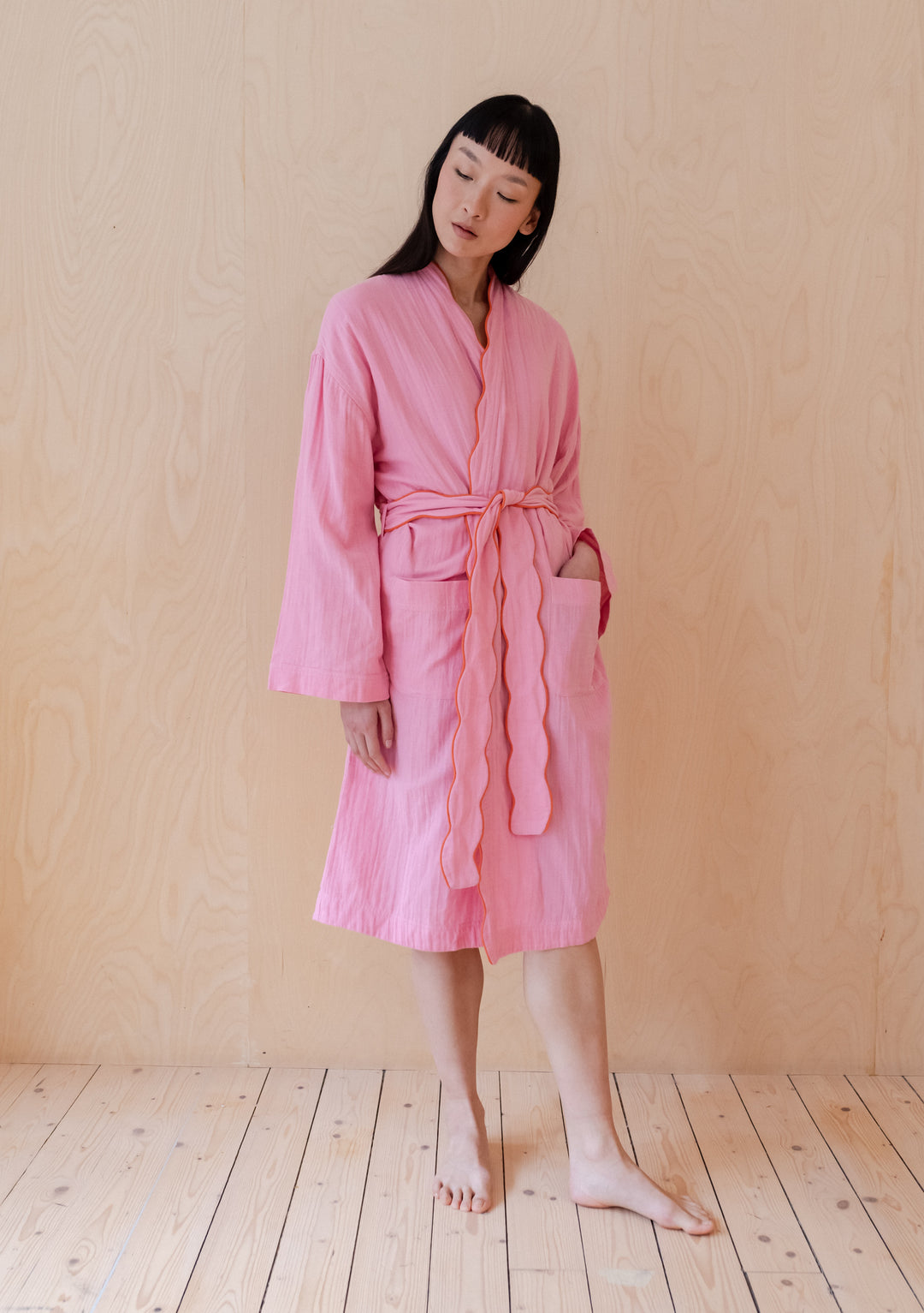 Pink cotton robe with scalloped edges from TBCo, shown against wooden backdrop, featuring tie waist and relaxed fit