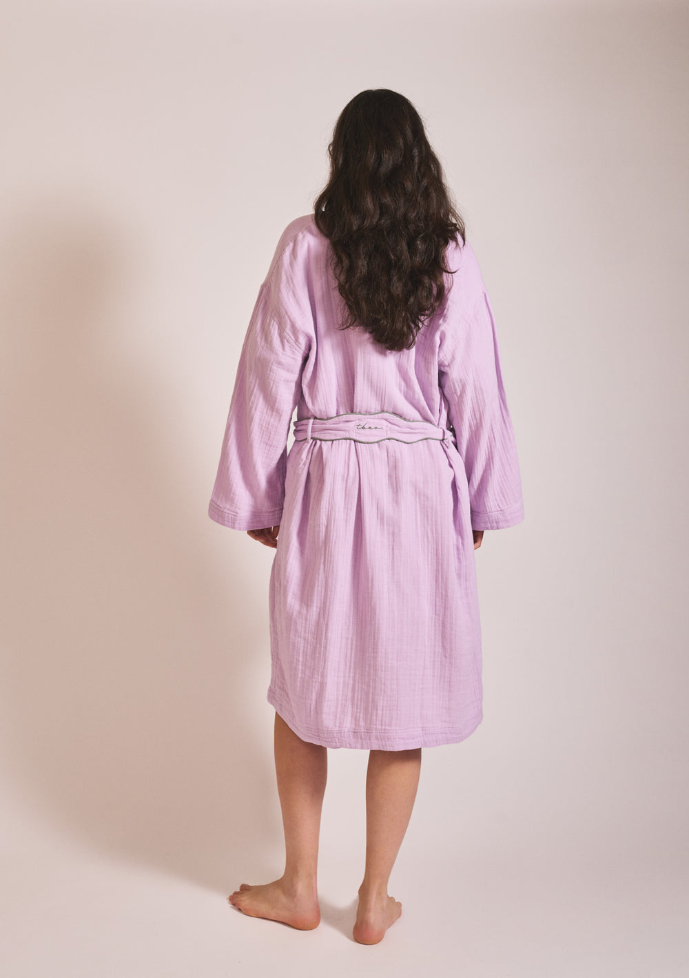 Portrait of a woman wearing TBCo Cotton Robe in Lilac, gallery image two showing back view with belt detail