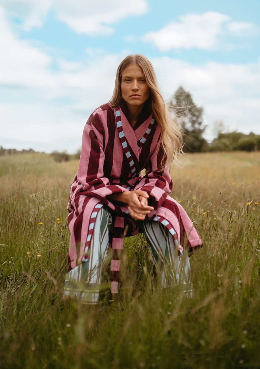 Woman wearing TBCo Cotton Bath Robe in Burgundy Stripe sitting in a grassy field under blue sky.