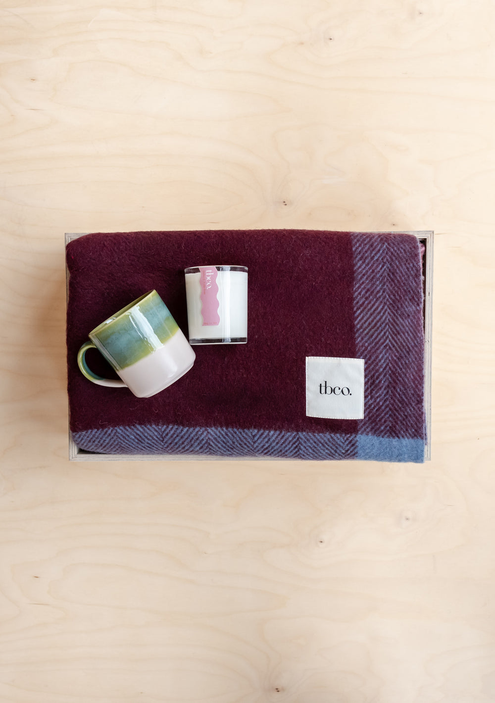 Ready-Made Cosy Blanket Gift Box by TBCo with berry check blanket, green dip mug, and scented candle on wood background