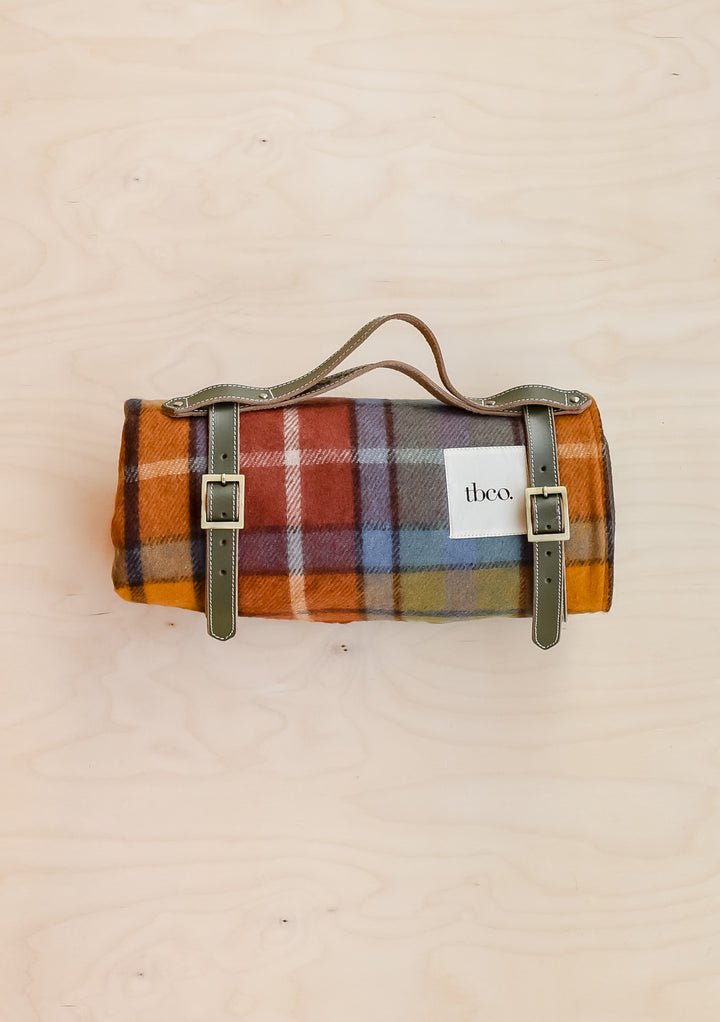 A TBCo recycled wool picnic blanket in Buchanan Antique Tartan, rolled up and held in a leather TBCo picnic carrier.