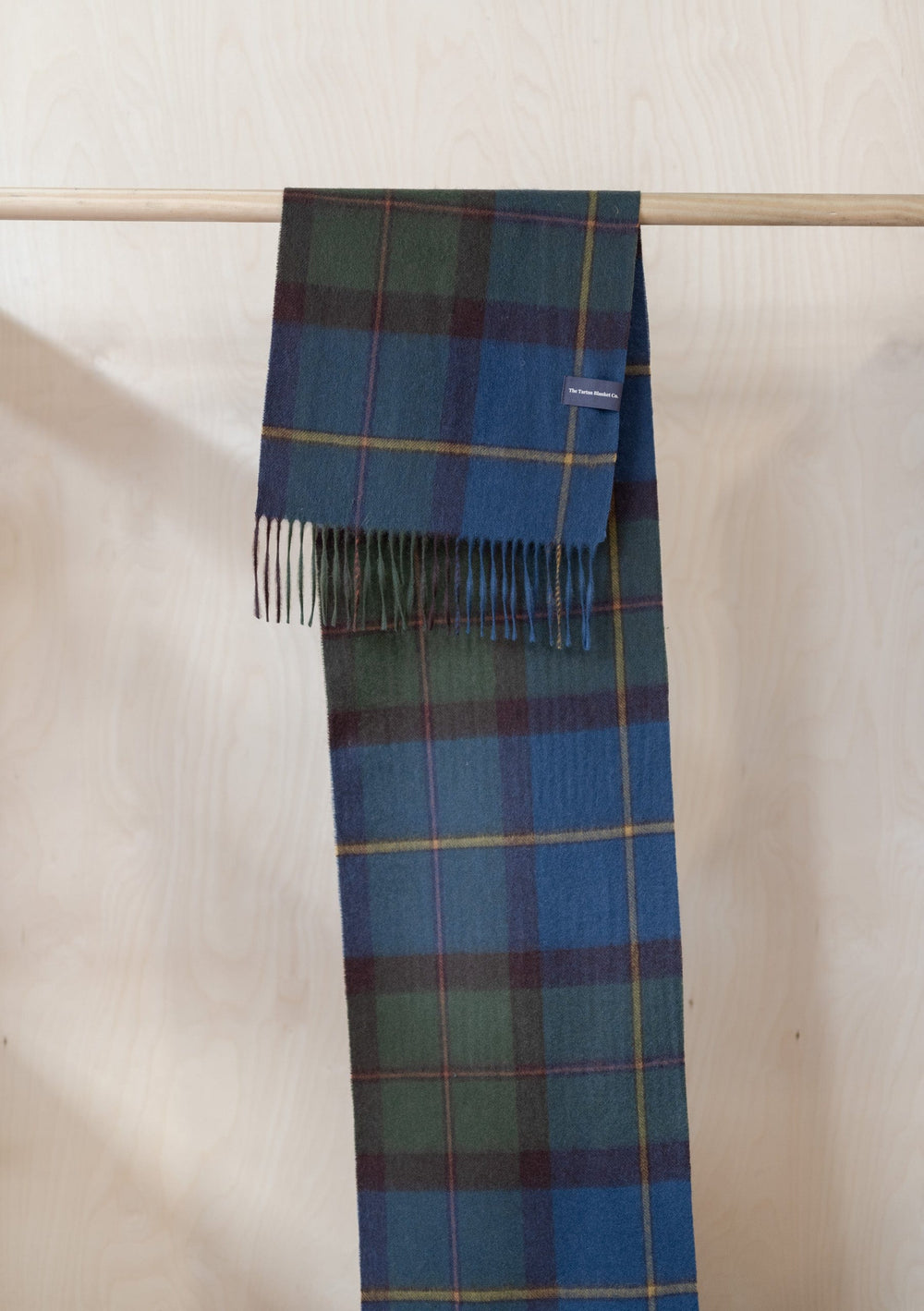 TBCo Macleod of Harris Antique Tartan Lambswool Scarf with fringe hanging on wooden rail, showcasing blue and green plaid pattern