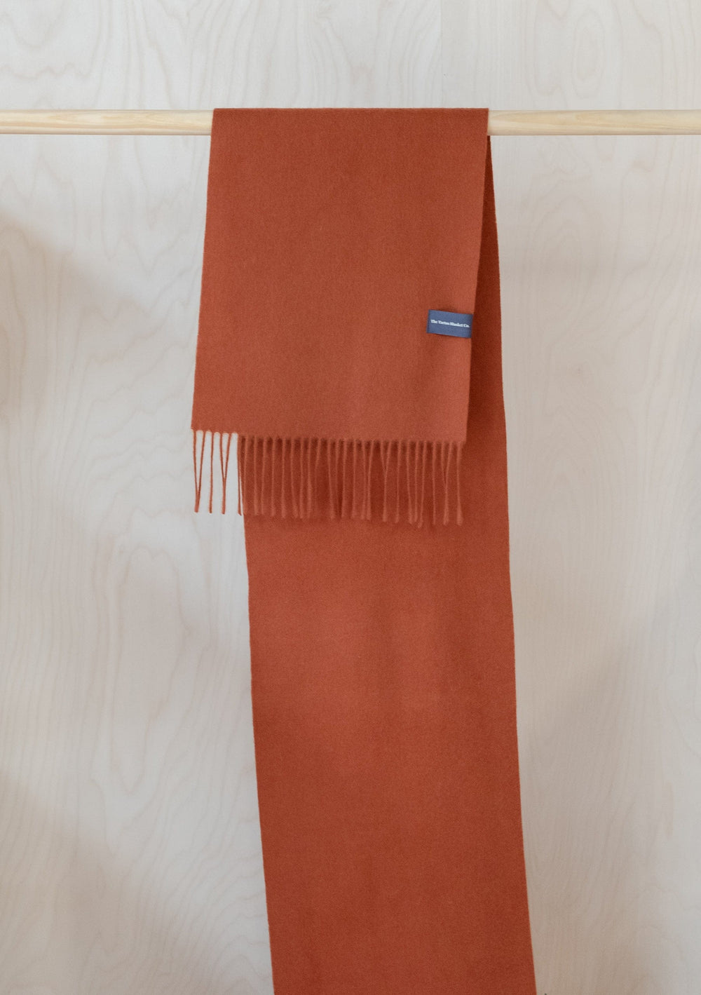 TBCo Men's Lambswool Scarf in Rust Melange hanging on wooden rail, showcasing fringed ends and luxurious woollen texture