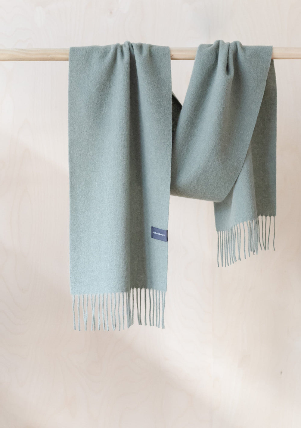 TBCo Lambswool Scarf in Sage Melange hanging on wooden rail, displaying soft texture and fringed ends in muted green colour