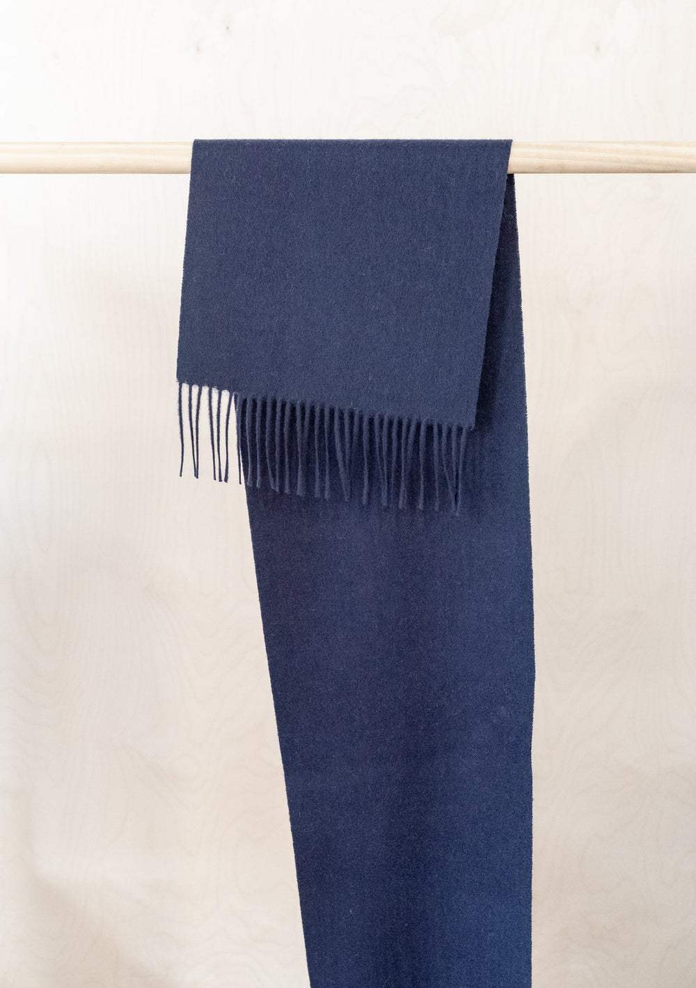 Soft navy blue TBCo lambswool scarf with fringed ends draped over wooden rail displaying classic winter accessory design